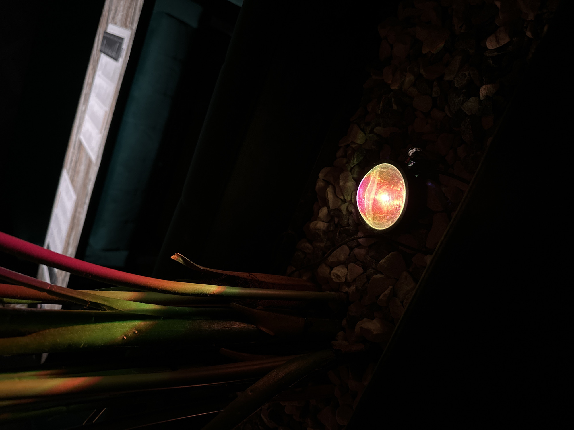 The image, taken at night in what appears to be an outdoor setting, features a round, illuminated light fixture nestled among small rocks. To the left of the light, a cluster of stems with red and green coloration is visible. In the upper left corner, a portion of a wooden structure with a light-colored surface is discernible, positioned next to a dark green, seemingly upholstered structure. The majority of the scene is cast in darkness, with the light fixture serving as the primary source of illumination. The location is specified as Chaikovsky, Russia.
