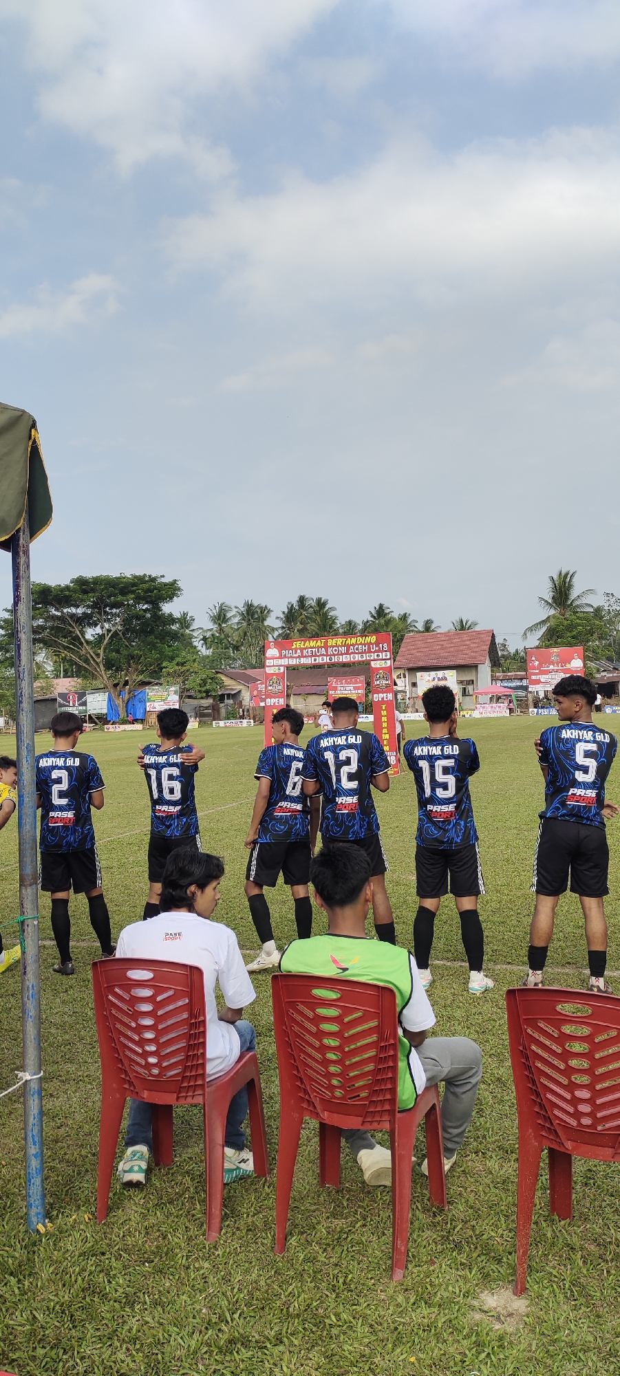 A group of young male soccer players, wearing matching blue and black jerseys with numbers on the back, are standing on a grassy field. The jerseys have the text "AKHYAR GLD" and "PASE SPORT" visible. The players are facing away from the camera. In the foreground, two young males are seated on red plastic chairs, also facing away from the camera. One is wearing a white t-shirt with "PASE SPORT" on it, and the other is wearing a green vest over a white shirt. Behind the players, a banner is displayed, reading "SELAMAT BERTANDING PIALA KETUA KONI ACEH U-13". Several palm trees and buildings are visible in the background, under a sky with scattered clouds. The setting appears to be an outdoor sports field in Kilometer Delapan, Indonesia.