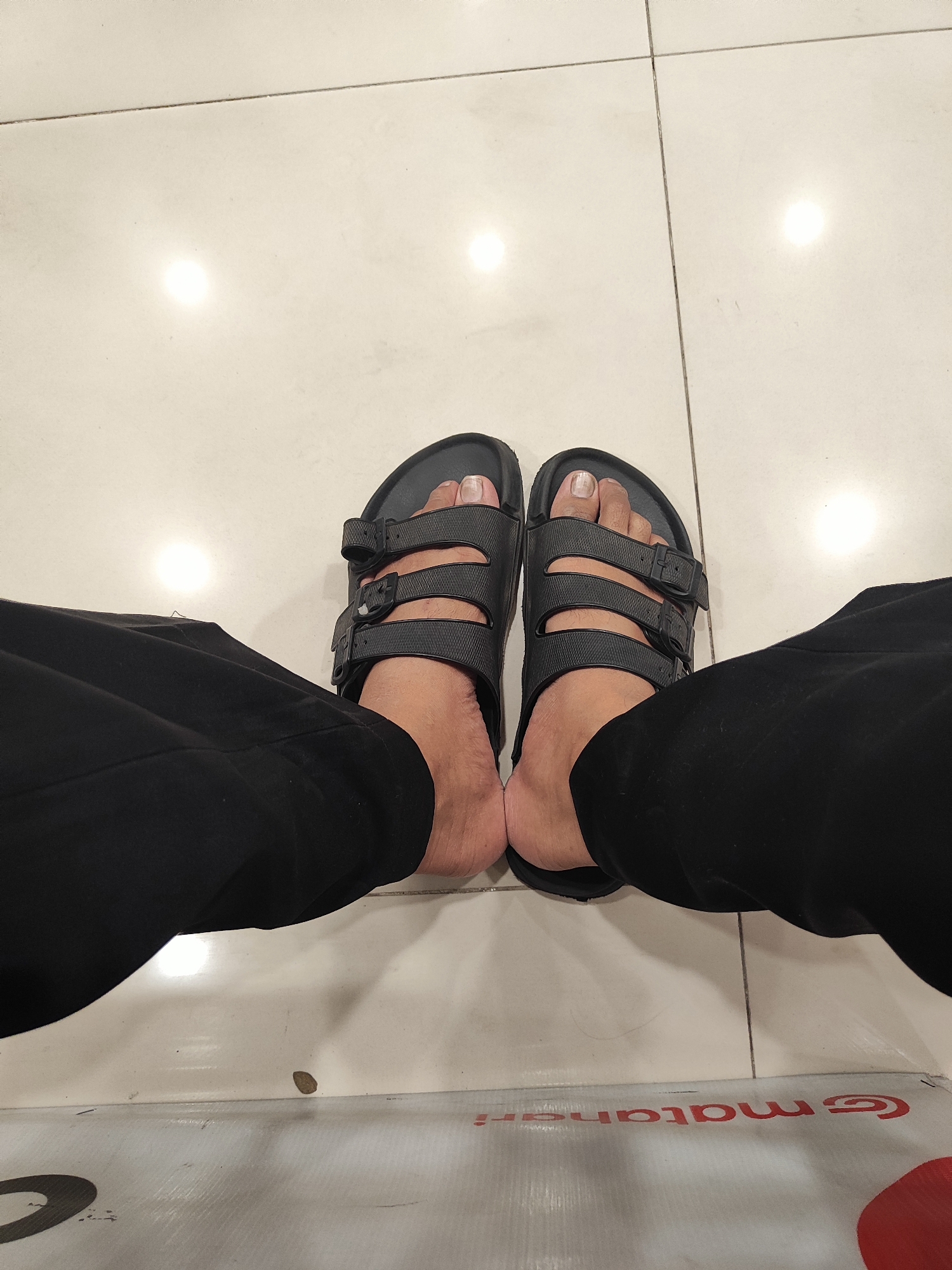 In Nangkaan, Indonesia, a person's feet are visible, clad in black two-strap sandals with adjustable buckles. The feet are positioned on a light-colored, tiled floor, with grout lines clearly demarcated. The person is wearing loose-fitting black pants. The overall environment appears to be indoors, possibly a public space given the flooring and lighting, which includes bright, circular reflections on the tiles. A portion of a large, white object with red lettering is visible at the bottom of the frame.