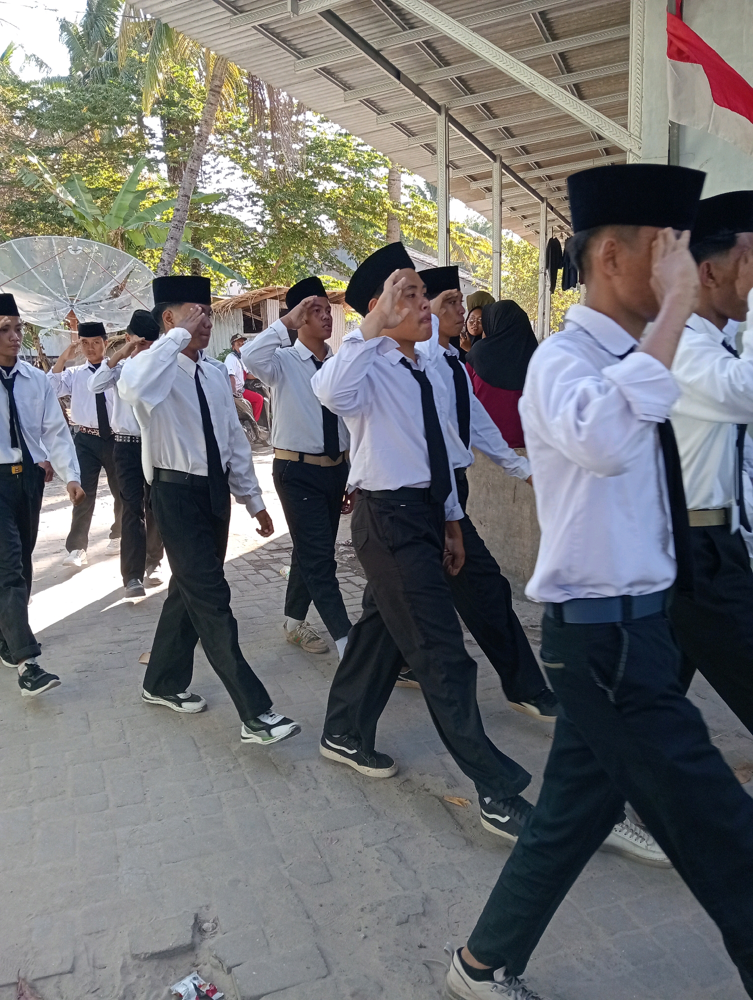 In Pagerungan Kecil, Indonesia, a group of young men in black peci hats and white long-sleeved shirts with black ties are marching in formation. They are wearing black pants and various types of sneakers. The men are saluting, with their right hands raised to their foreheads. The procession is taking place outdoors on a paved walkway. In the background, there are palm trees, a satellite dish, and a building with a corrugated metal roof. A red and white flag, likely the Indonesian flag, is visible in the upper right corner. The sun is casting shadows, suggesting it is daytime.