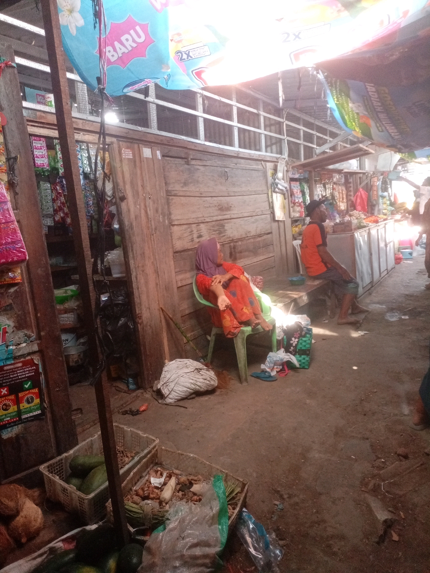 The image depicts a street market scene in Sluke, Indonesia. In the foreground, a woman wearing a purple hijab and an orange and red patterned dress is seated on a green plastic chair. Beside her, on the ground, are several items including a white woven bag, a shopping bag with a floral pattern, and a pair of blue sandals. To the left of the woman, there are two plastic baskets. One contains several green fruits, possibly avocados, and the other contains roots and spices, along with some green leaves. Further to the left, a wooden structure is visible, with shelves holding various packaged goods.
In the midground, a man wearing an orange t-shirt and dark shorts is sitting on a wooden bench. He is facing away from the camera, looking towards the right. Behind him, there appears to be a market stall with goods displayed on shelves and a counter.
The environment is rustic, with wooden structures and a dirt ground. The market is covered by tarpaulins and fabric awnings, suggesting it is outdoors or partially enclosed. The lighting appears to be natural sunlight, with some areas casting strong shadows. The overall impression is that of a busy, informal marketplace.