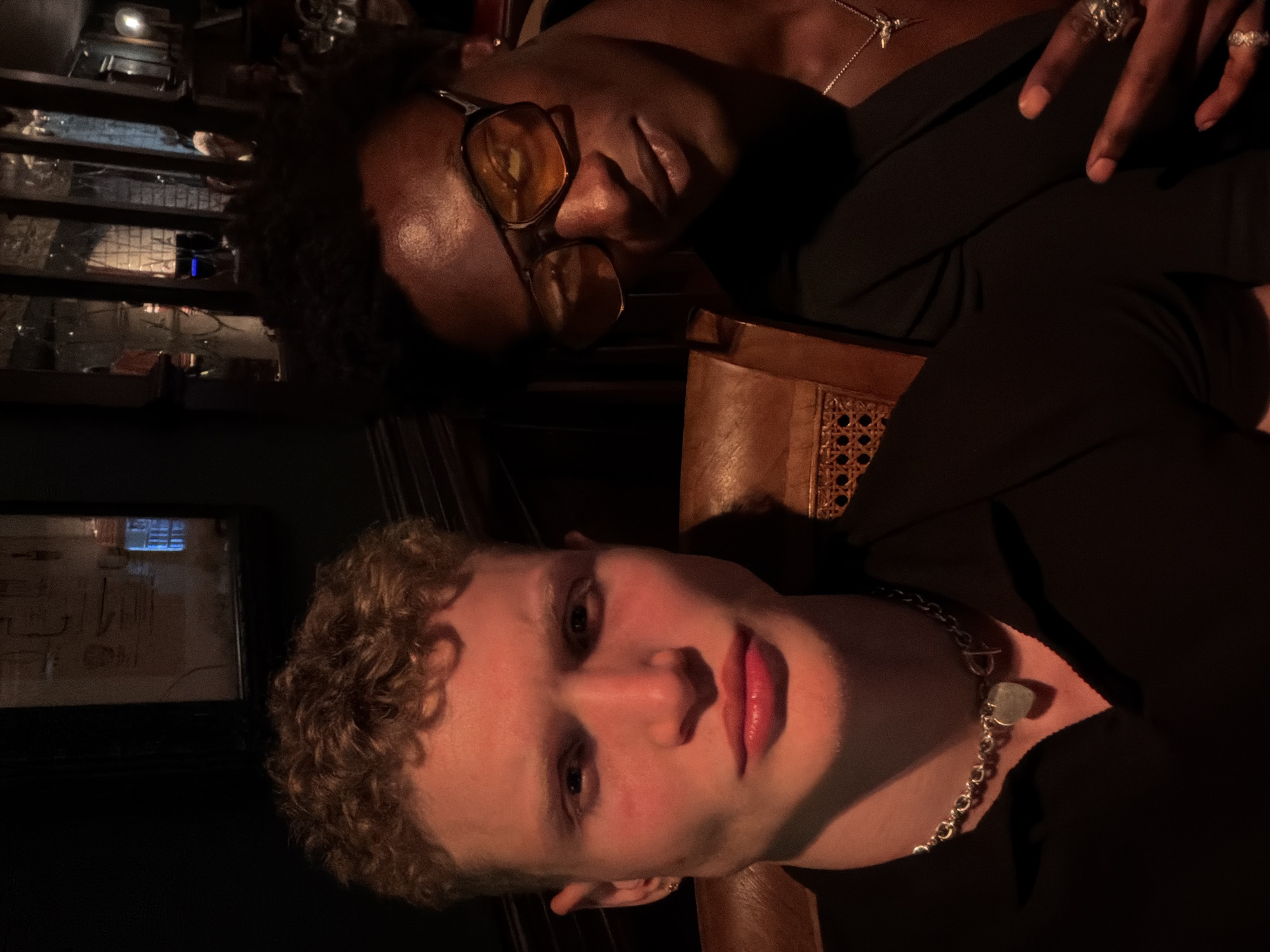 Two individuals are captured in a dimly lit, intimate indoor setting, possibly a bar or restaurant in London, United Kingdom. The image is oriented vertically (portrait mode), featuring one person prominently in the foreground and another slightly behind and above.
The person in the foreground has fair skin, light brown curly hair, and is looking directly at the camera with a neutral expression. They wear a thick silver chain necklace with a light, possibly opalescent, square-shaped pendant. Their lips are full, appearing to have gloss or light lipstick, and are highlighted by a warm, reddish-orange light source coming from the right.
Behind and slightly to the left of the first person is a second individual with dark skin and dark curly hair. They are wearing amber-tinted sunglasses, which obscure their eyes, and a thin silver necklace with a small, bird-like or abstract pendant. Their fingers are adorned with rings, including an ornate ring on the index finger and another on the ring finger. This person looks towards the lower right, with a relaxed posture.
The background reveals elements of the venue. To the left of the second person, there are multi-tiered display shelves or cabinets with glass fronts, reflecting various objects and light sources, including a distinct blue light on one shelf. To the left of the first person's head, a dark framed object, possibly a mirror or picture, reflects subtle patterns and light. A wooden chair with a cane back is visible in the middle ground, adding to the classic ambiance.
The lighting is low and atmospheric, suggesting an evening or nighttime setting. Direct warm light illuminates parts of both individuals' faces, creating shadows and highlighting facial features. No visible text is discernible in the image.