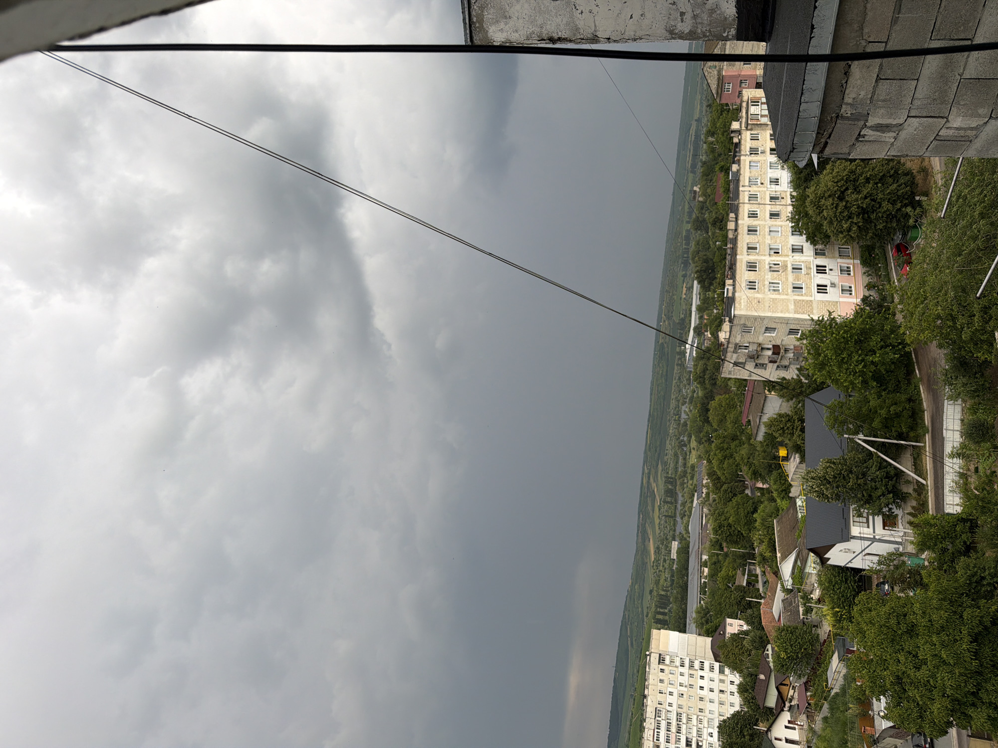 The image depicts an overcast sky with heavy grey clouds over a residential area in Strășeni, Moldova. In the foreground, the edge of a building constructed with concrete blocks is visible on the right side. A thin wire runs diagonally across the frame from the upper left to the lower right. Below the clouds and in the midground, several buildings are visible, including a large, light-colored apartment building with many windows. Surrounding the apartment building are smaller houses with various roof colors and numerous trees. The landscape beyond the residential area consists of rolling green hills and fields, with a body of water possibly visible in the distance. The overall scene suggests a slightly gloomy or pre-storm atmosphere. No people or specific actions are clearly discernible.