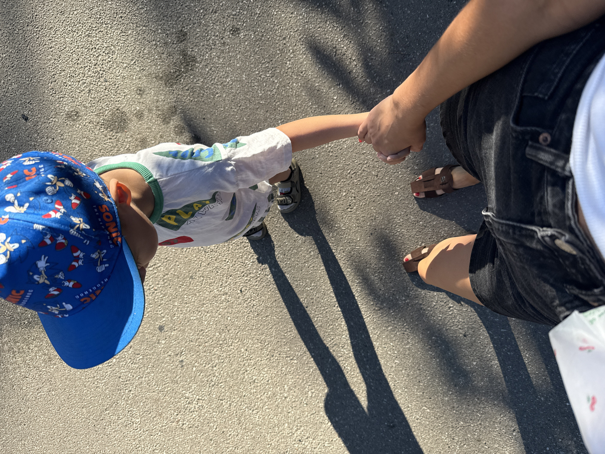 In an outdoor urban setting, possibly in Kyiv, Ukraine, a child wearing a blue Sonic the Hedgehog-themed baseball cap and a white t-shirt with a green collar is walking while holding hands with an adult. The child is also wearing grey sandals. The adult is wearing black denim shorts, a white t-shirt, and brown sandals with red nail polish visible on their toes. They are walking on a paved surface, likely a sidewalk or street, as indicated by the asphalt texture and the long shadows cast by the sun. The lighting suggests it is daytime.