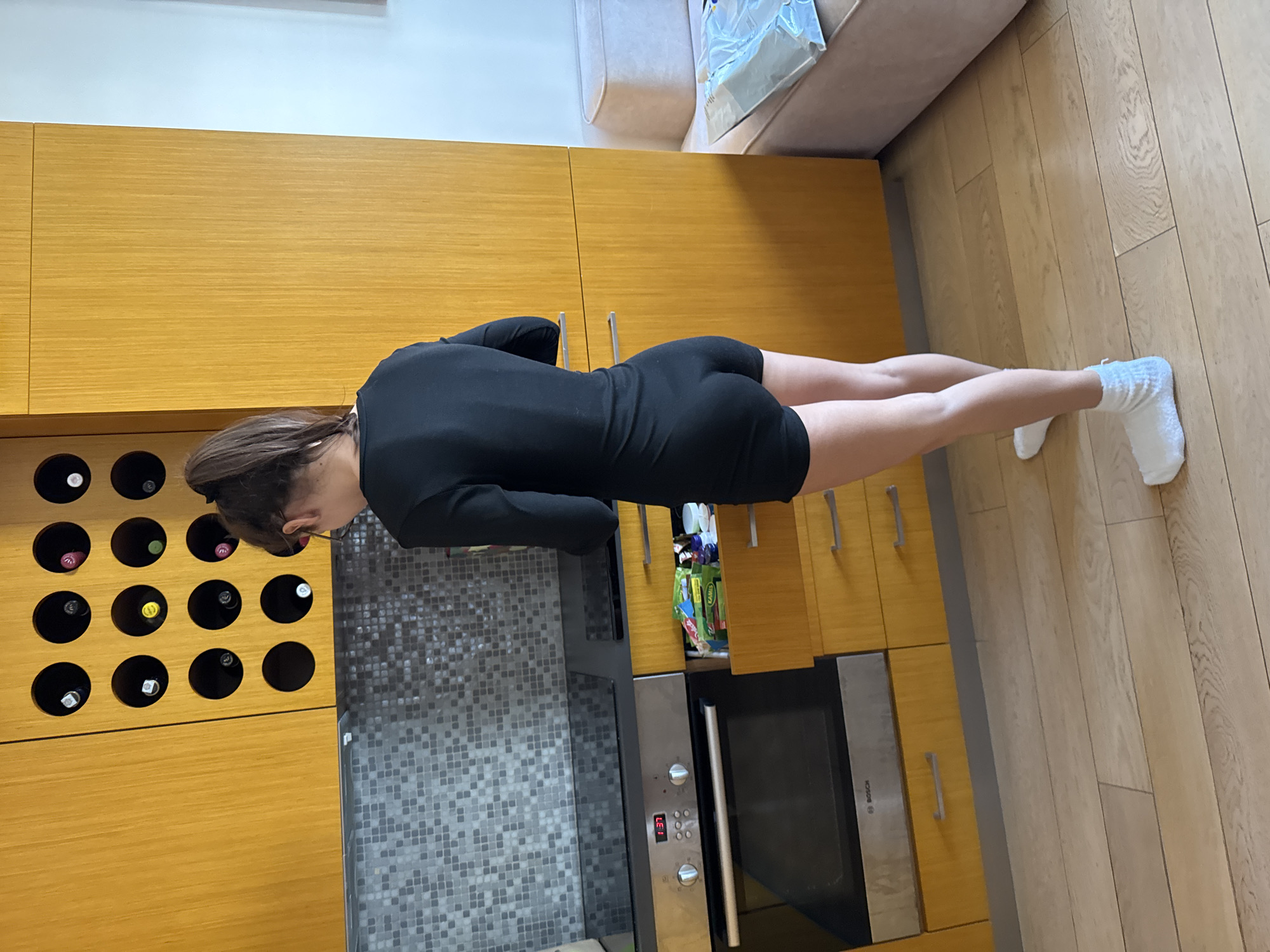 The image, rotated 90 degrees counter-clockwise, captures an indoor scene featuring a person in a modern kitchen, consistent with the specified location of Warsaw, Poland.
The main subject is a person, likely female, shown from the back in a body-extended pose, suggesting either an exercise like a plank or reaching for something low. She is wearing a black long-sleeved bodysuit or short jumpsuit and white, fluffy socks. Her head is bowed, looking downwards.
The setting is a well-lit kitchen characterized by light-colored, honey-toned wooden cabinetry. One section of the cabinetry features an integrated wine rack holding several bottles of wine. A gray, square-tiled backsplash is visible behind the person. An integrated Bosch oven or microwave is built into the cabinets, displaying "1:37" on its digital clock and the text "LEI" below the time. Above this appliance, a cabinet door is slightly ajar, revealing colorful packaging inside, including items branded "Kamis," a known Polish food brand, which strongly supports the Warsaw location context.
The floor is covered with light-colored wooden floorboards. In the background, a light-colored sofa and a plain wall are visible, indicating an open-plan living area.
The time of day, suggested by the appliance clock, is 1:37, though whether it is AM or PM is not discernible from the indoor lighting. No specific weather conditions or emotions are evident.