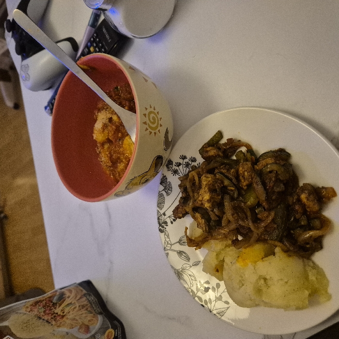 A bowl of corn with a spoon sits beside a plate of dark, sauteed vegetables and mashed potatoes. The bowl has a red interior and patterned exterior with a sun design. The plate is white with a floral pattern around the edge. A partially visible package of food is near the bottom of the image, and a game controller sits near the bowl. A container of liquid, possibly milk, and an object are behind the controller. The setting is a flat, white surface.