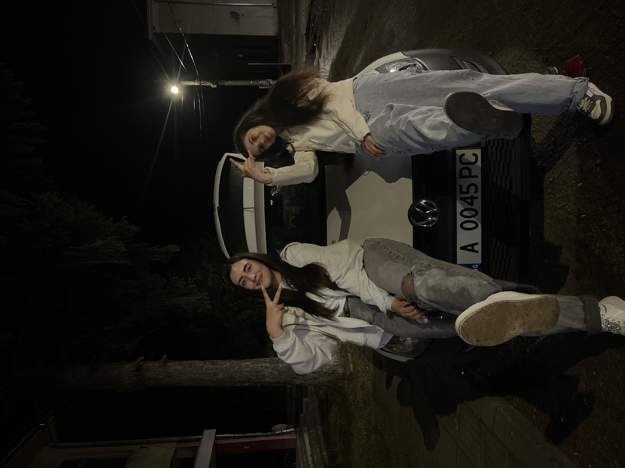 Two young women are reclining on the hood of a car, posing for a photo. The car is white and has a license plate that reads "A 0045 PC." The scene appears to be at night, with artificial lighting illuminating the area. Trees are visible in the background, and there are buildings nearby. One woman is wearing a light-colored jacket and light-wash jeans, while the other is wearing a gray sweatshirt and gray jeans. The location is Lozarevo, Bulgaria.