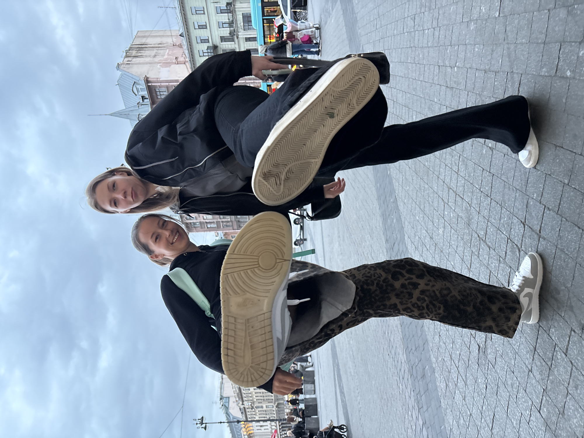 Two young women are posing in a city square on a cloudy day. The woman in the foreground is wearing a black hoodie, a light green backpack, and leopard print pants. Her left foot is planted on the ground, while her right leg is extended towards the camera, showing the sole of a white sneaker. She is smiling at the camera. The woman in the background is wearing a black jacket and pants. Her left foot is planted on the ground, while her right leg is extended towards the camera, showing the sole of a white sneaker. She is looking at the camera. In the background, there are buildings, people, and buses. The location is Saint Petersburg, Russia.