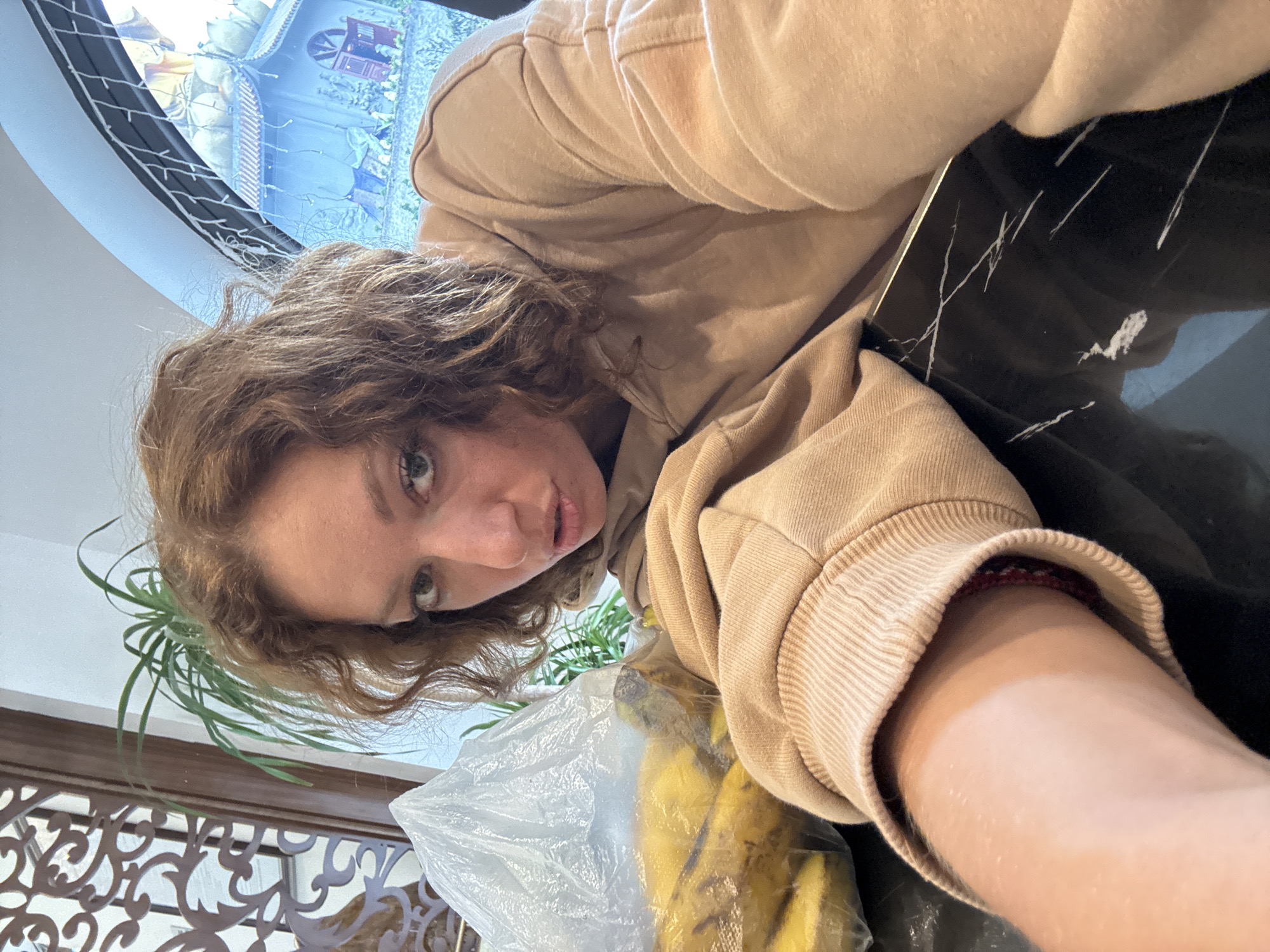 A woman with curly auburn hair and fair skin is positioned at an angle in the image, wearing a tan hoodie. A bunch of bananas in a plastic bag are near her. A black marble surface is in the background, along with a decorative window displaying a building reminiscent of East Asian architecture. A potted plant is also visible in the lower-left corner.
It is not possible to determine the specific location in Binh Lu, Vietnam based on the provided image alone.