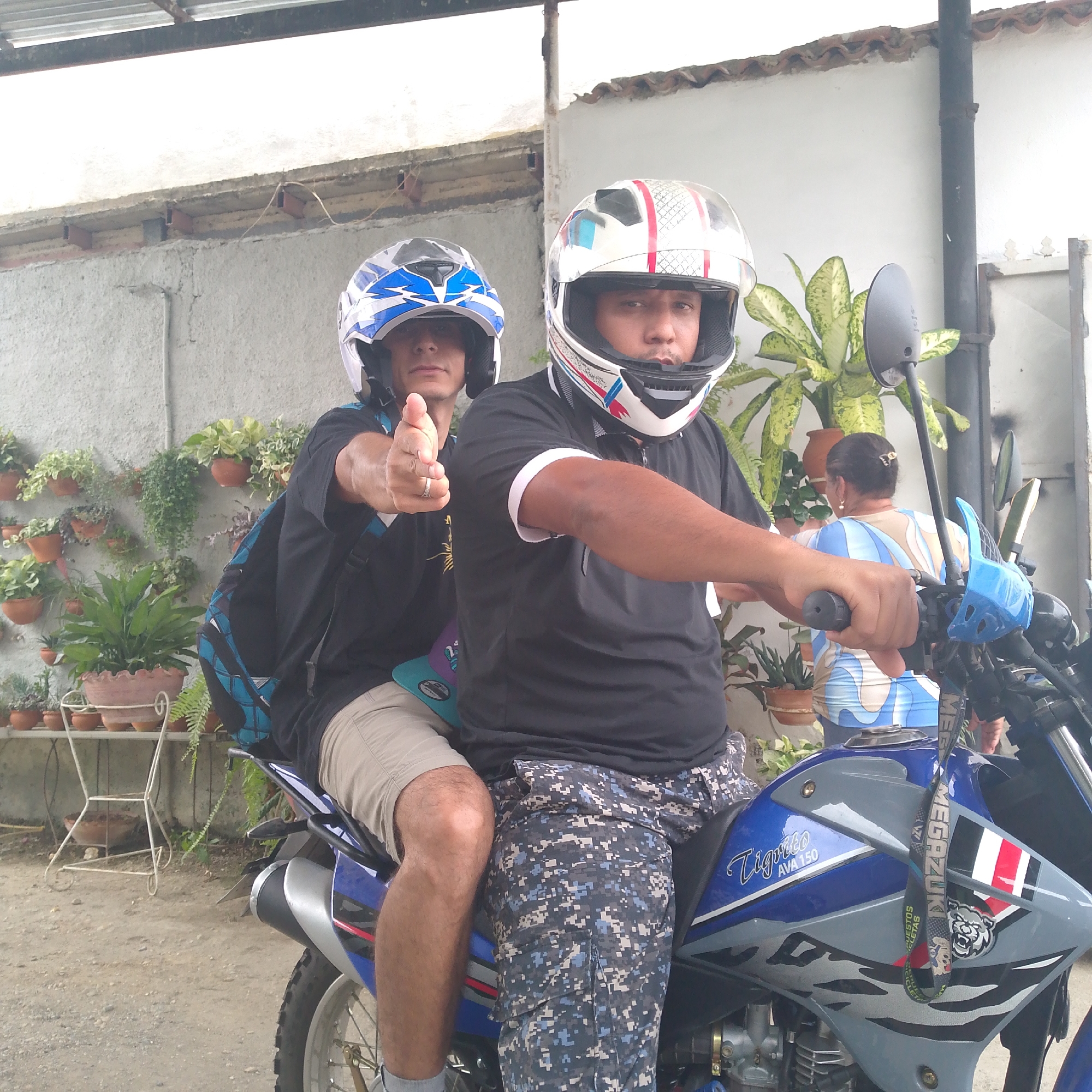 Two men are depicted sitting on a blue and grey motorcycle in an outdoor setting in Tinaquillo, Venezuela, during daylight hours. The man in the foreground, wearing a white helmet with red and blue details, holds the handlebars and looks forward. He is dressed in a black short-sleeved shirt and blue digital camouflage trousers.
Behind him sits another man, wearing a white and blue helmet, who is actively pointing directly at the viewer with his right hand. He wears a black t-shirt, light khaki shorts, and has a blue and black backpack. The motorcycle itself features distinct blue and grey panels.
The background consists of a textured concrete wall, which is extensively decorated with a variety of potted plants and hanging greenery, suggesting a domestic garden or patio area. Above, a corrugated metal roof structure is visible. To the far right, partially obscured by plants and the motorcycle, a woman with her back to the camera can be seen wearing a blue and white patterned top.
Visible text on the motorcycle's side reads "Tigrillo AVA 150." A lanyard hanging from the handlebars also displays text, including "MEGADZUKII" and "PUESTOS BICICLETAS." The scene captures a casual moment, possibly before or after a ride.