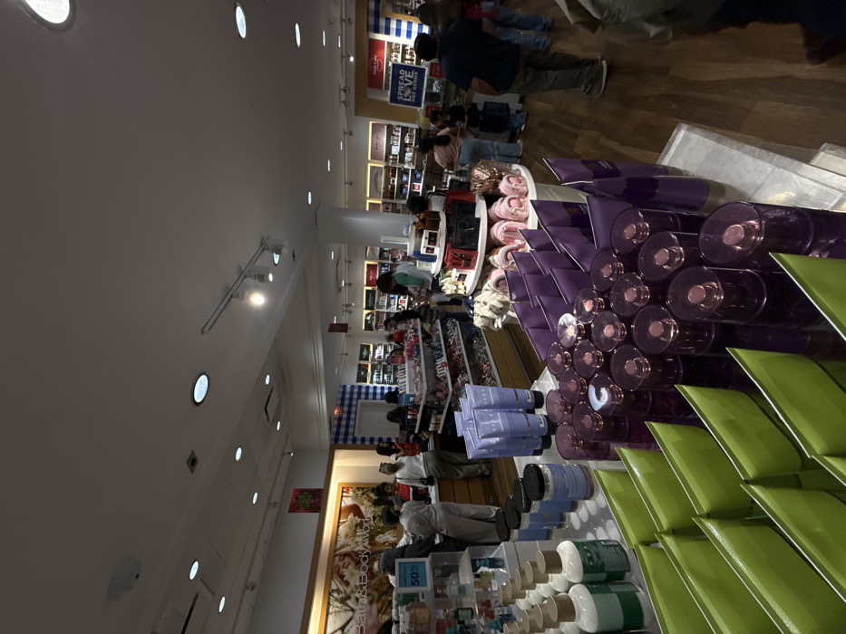 Inside a brightly lit Bath & Body Works store, customers browse merchandise. Shelves display candles, lotions, and other fragrant items. A large display features purple and green products, creating a visually appealing arrangement. People can be seen shopping in the aisles, adding to the busy atmosphere of the retail space.