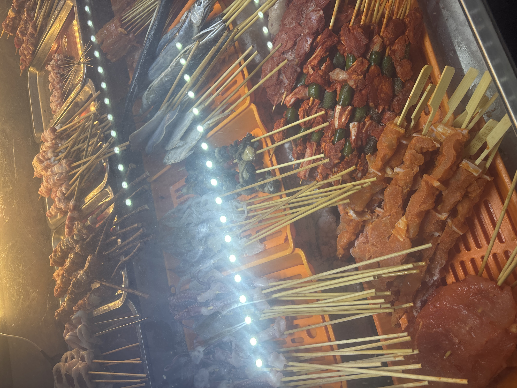 This image depicts a display of various skewered raw food items, likely in a food stall or market in Suzhou Industrial Park, China. The items are arranged on multiple levels, illuminated by overhead lighting which causes some glare.
In the foreground and middle ground, various types of meat skewers are visible. These include what appears to be marinated red meat, some with green pepper pieces interspersed, and others with pieces of what looks like chicken or pork. Some skewers are densely packed with meat, while others have larger, more distinct cuts.
Further back and on upper levels, other food items are displayed. There are skewers of what appear to be seafood, including what could be squid or octopus tentacles, and several whole fish are laid out on skewers. There are also some darker, possibly mushroom or vegetable skewers visible.
The food is presented in plastic trays and on metal trays. The overall environment suggests a commercial setting focused on selling raw ingredients for grilling or other cooking methods. There are no people visible in the image, and the focus is solely on the food display. The lighting creates a bright, somewhat reflective surface over the food.