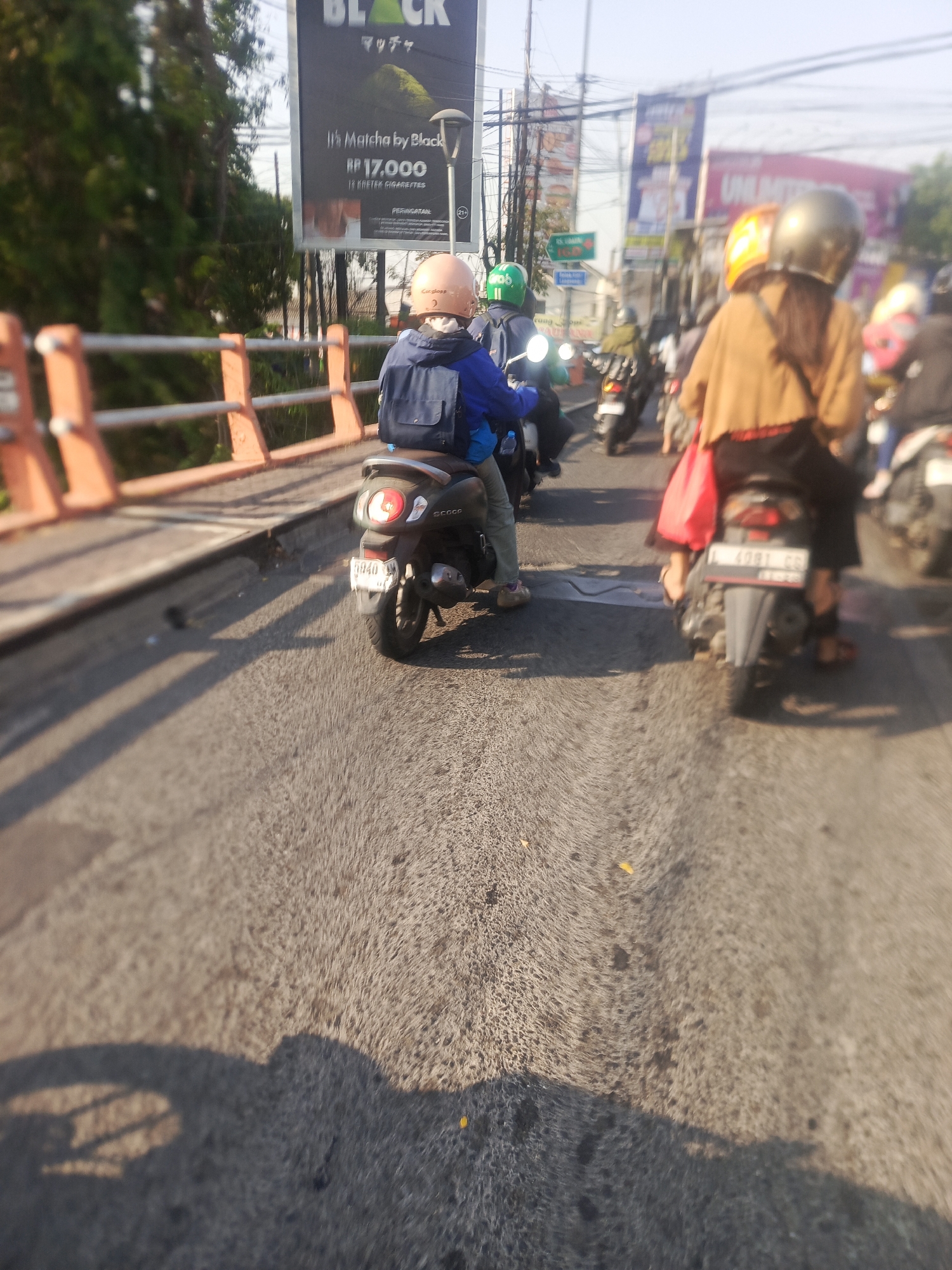 This is a photograph taken on a sunny day, likely during the day, from the perspective of someone riding on the back of a motorbike. The scene depicts a traffic jam on a bridge in Surabaya, Indonesia, as evidenced by the billboard in the background advertising "Black Matcha" with the price in Indonesian Rupiah (Rp 17,000) and the license plate prefixes common in East Java.
In the foreground, a dark-colored scooter is stationary, with its rear tire and license plate visible. The rider of this scooter is wearing a blue jacket and an orange helmet, with a backpack on their back. The scooter's tail light is illuminated. The scooter is positioned to the left of the frame.
To the right of the frame, another scooter is slightly ahead. The rider on this scooter wears a brown outer garment and a silver helmet. A bright red bag is slung over their shoulder and rests on the back of the scooter. The scooter's rear tire and license plate are also visible.
Several other motorbikes are visible in the middle ground, forming a queue. Some riders are wearing helmets of various colors, including green. The road surface appears to be asphalt, and the sun is casting long shadows. In the background, there are trees, power lines, and several billboards, including one for "Black Matcha" with Japanese text. The bridge railing, painted orange, runs along the left side of the frame. The overall impression is one of heavy urban traffic on a bright day.