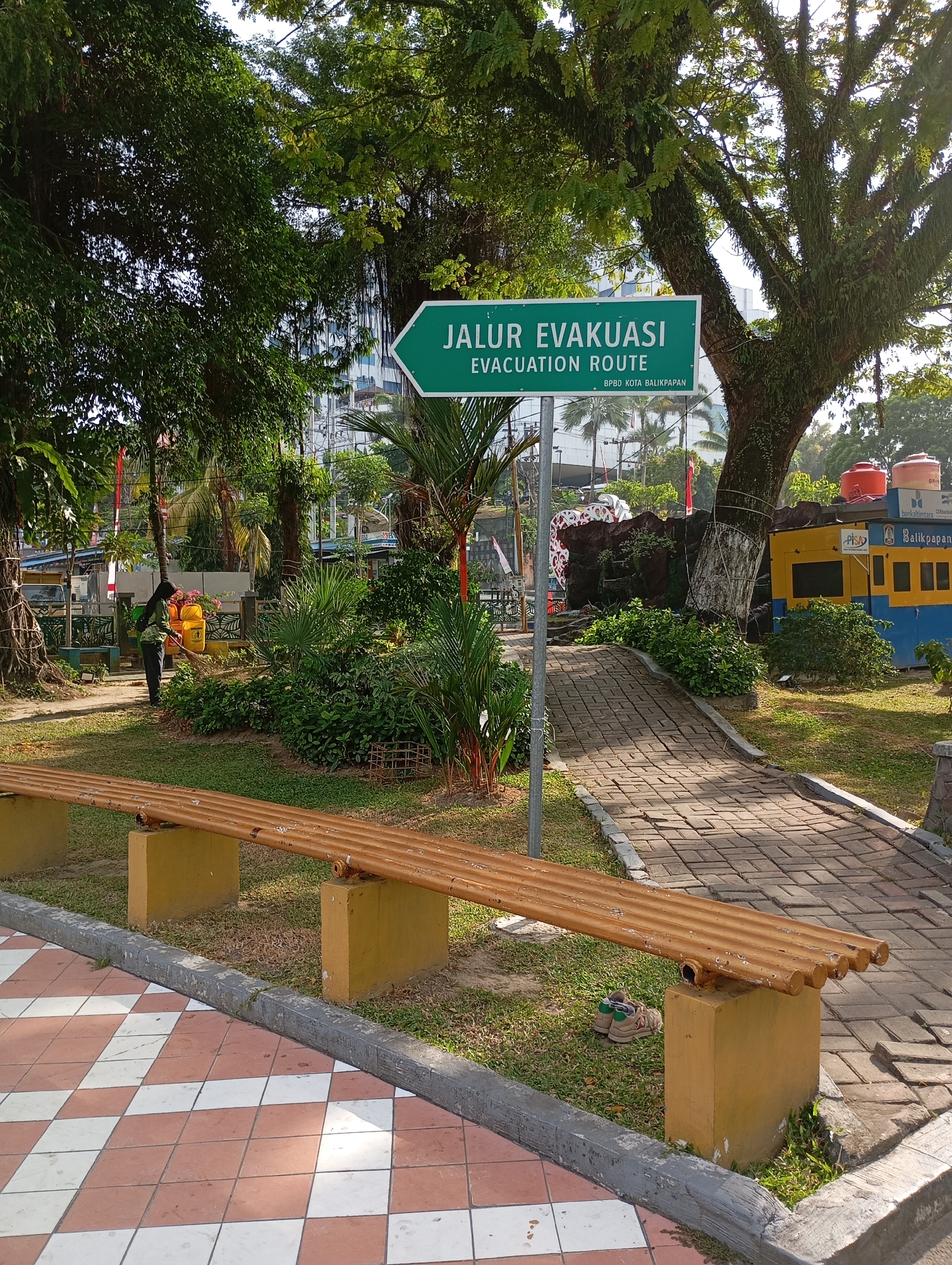 The image depicts an outdoor scene in Balikpapan, Indonesia, likely a park or public area. A prominent green sign with white text reads "JALUR EVAKUASI EVACUATION ROUTE" and below that, "BPBD KOTA BALIKPAPAN," indicating an evacuation route managed by the Balikpapan City Regional Disaster Management Agency. The sign is positioned near a large, old tree with extensive foliage.
In the foreground, a long, wooden bench with a weathered, corrugated surface rests on yellow concrete supports. The bench is situated on a grassy area next to a paved walkway made of interlocking brick pavers in a diamond pattern. A pair of beige shoes with green accents are visible under the bench.
To the left of the bench, a person, appearing to be a woman with long dark hair, is sweeping the ground with a broom. She is wearing a green shirt and dark pants. Beside her are two large yellow buckets. Behind her, a low fence and some plants are visible, with Indonesian flags displayed.
In the mid-ground, the paved walkway curves and leads towards a decorative rock structure. Behind this structure is a brightly colored, modular building painted yellow and blue. Red and orange water tanks are visible on top of this building. The background reveals more trees and buildings, suggesting an urban or semi-urban environment. The overall scene is bathed in sunlight, indicating daytime.