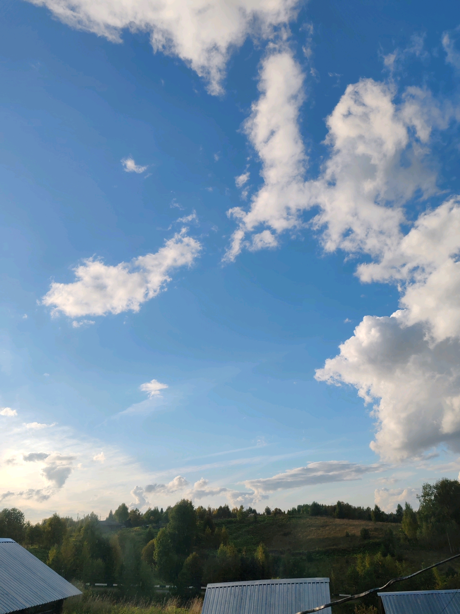 The image depicts a rural landscape with a vast blue sky filled with scattered white cumulus clouds. The horizon is occupied by a gently rolling hill covered in dense green trees, suggesting a forested area. The foreground features the metal roofs of two structures, likely rural houses or sheds, positioned to the left and right edges of the frame. There is no indication of human presence or any activity within the scene. The overall impression is one of a tranquil, natural setting. Based on the context provided about the city of Чухлэм, Russia, this scene could be representative of the countryside surrounding such a location.