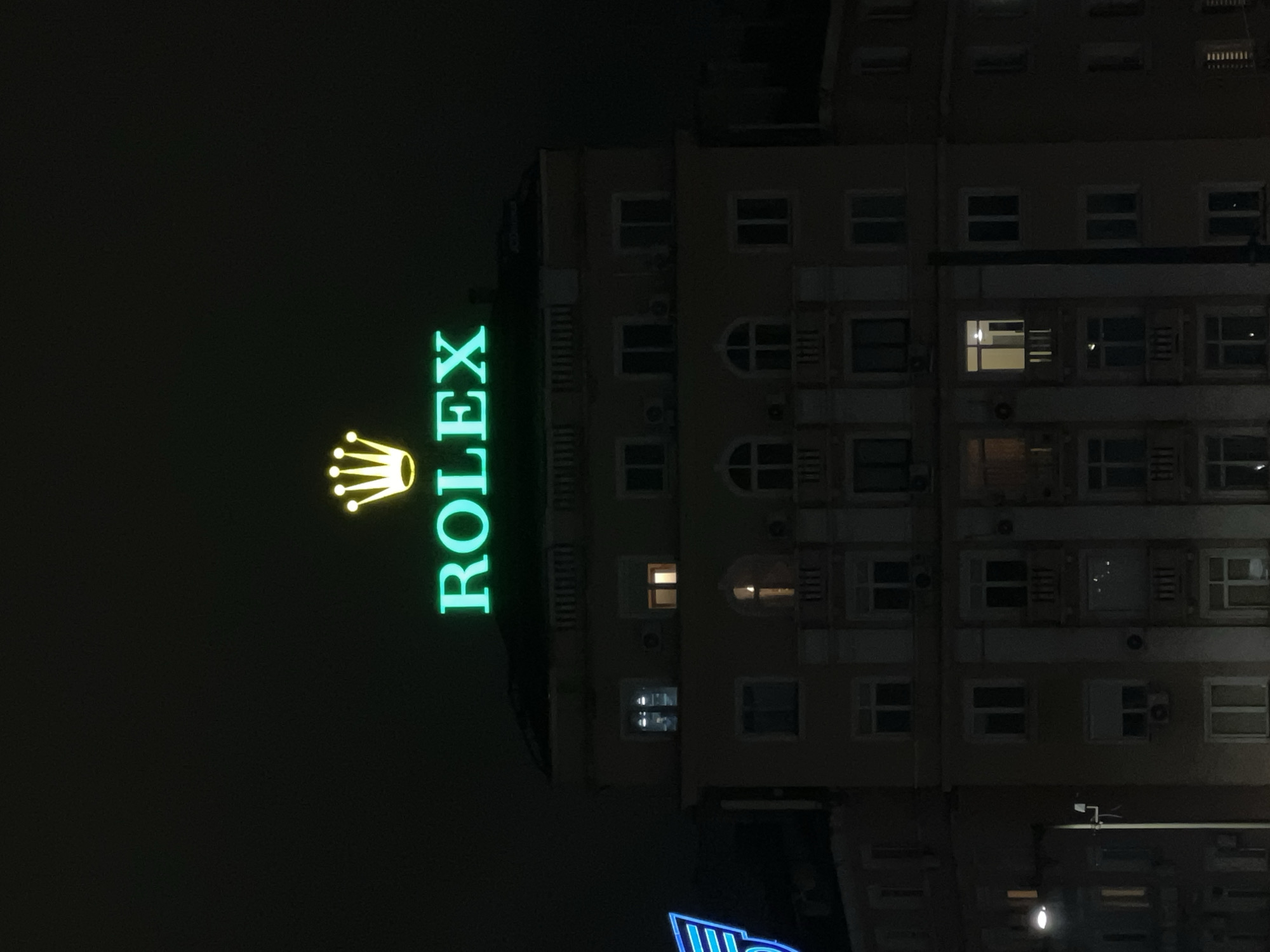 The image captures a multi-story, light-colored building at night, located at Vitosha Blvd 2, Sofia, Bulgaria. Prominently positioned on the rooftop is an illuminated Rolex sign, displaying the brand name in bright green neon letters and its distinct crown logo in yellow neon above. The building's facade features numerous windows across multiple floors, some of which exhibit internal illumination. Architectural details include decorative window frames, small balconies, and visible air conditioning units mounted below several windows. To the left of the main building, a partial blue neon sign with linear, possibly wing-like, elements is visible. A streetlamp in the lower-left foreground provides limited ground-level illumination. The sky is dark, indicating a nighttime setting, and no individuals are discernibly present within the frame.