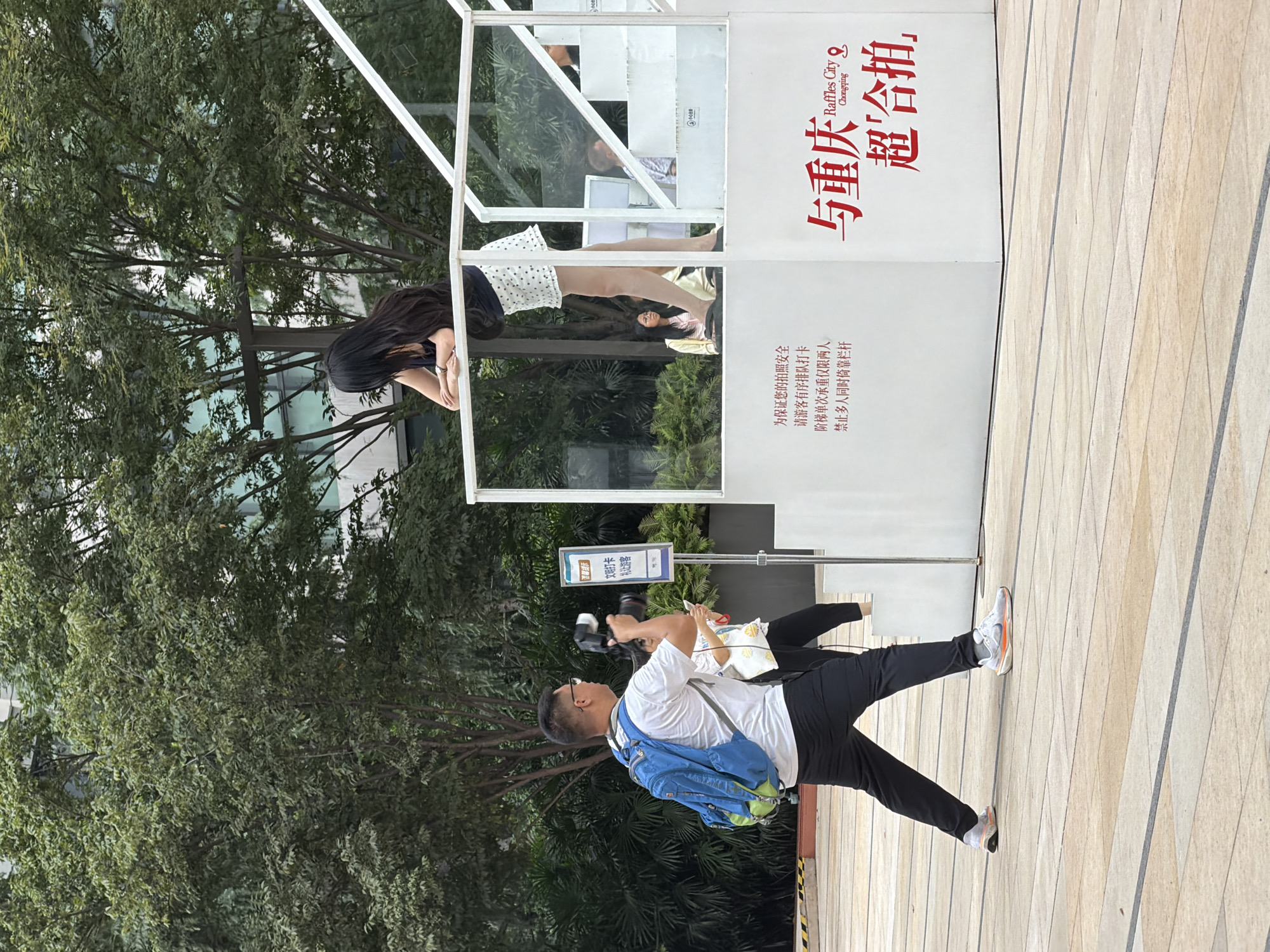 The image depicts an outdoor scene in the Yuzhong District of China, likely at Raffles City Chongqing, given the text on the building. A man with a blue backpack is in the foreground, holding a camera and appearing to take a picture of a woman leaning over a railing on an elevated platform. The woman has long black hair and is wearing a dark top and a white polka-dot skirt. The platform has white stairs and glass barriers. A sign is visible on the platform with Chinese characters. The ground is paved with light-colored tiles. Trees and a building are visible in the background. Another person can be seen behind the man with the camera, and a few individuals are visible on the stairs of the platform.
