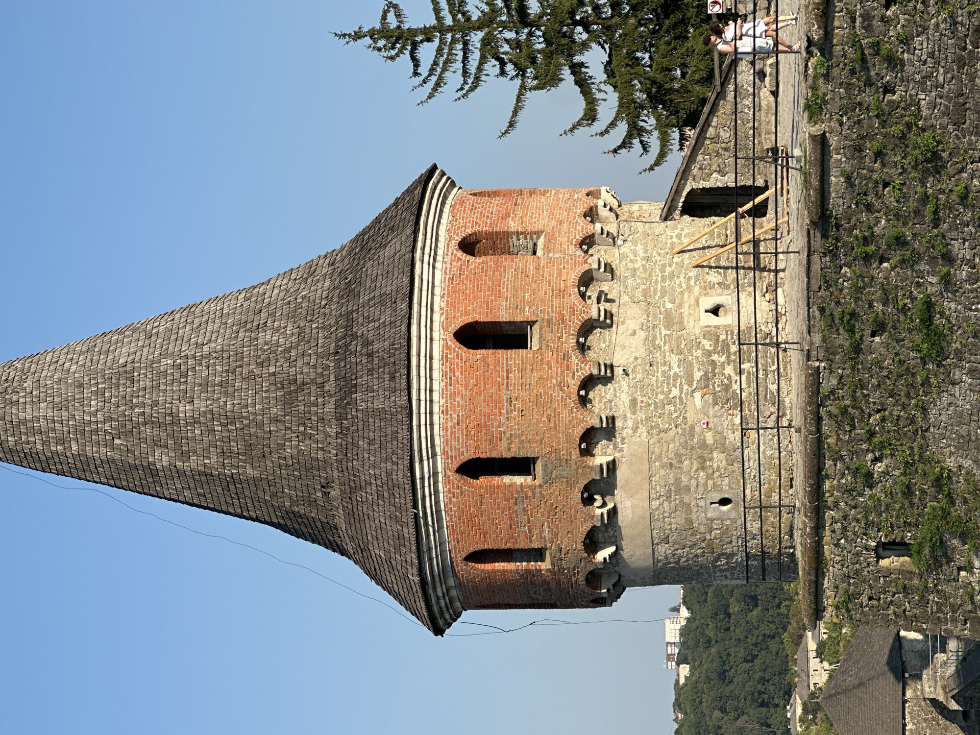 A close-up, slightly elevated view captures the upper portion of a circular stone and brick tower with a wooden shingled conical roof. The tower has a rough stone base, transitioning to red brick in its upper section, punctuated by arched windows. Decorative stone arches support the brickwork. A dark wire dangles from the roof's edge. A tall evergreen tree stands to the right, partially obscuring the sky. In the middle ground, a stone walkway with a black metal railing leads to a wooden doorway in the tower's side. Two individuals, a man and a woman, walk along the railing. The background reveals a lush green hillside with buildings and a clear blue sky.