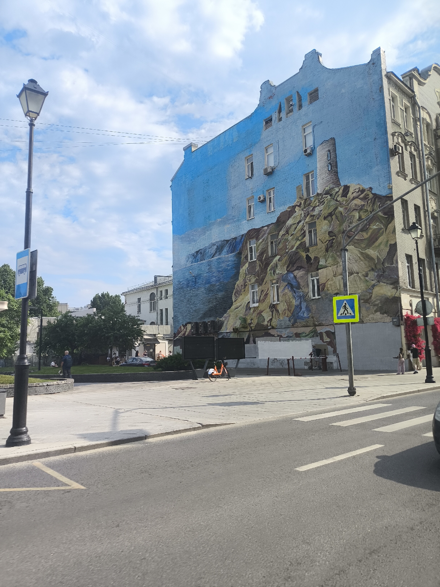 The image depicts a street scene in Moscow, Russia, featuring a large building with a mural painted on its facade. The mural depicts a landscape with a waterfall and rocky terrain, incorporating the windows of the building into the artwork. The sky above the mural is painted blue.
To the left of the mural is a lamppost with a bus stop sign attached. A paved area stretches from the building to a street with a pedestrian crosswalk. Several people are present in the background, some sitting on a bench and others walking. A pedestrian crossing sign is visible in front of the building. There is a car on the far right in the street. The weather appears to be sunny with scattered clouds.