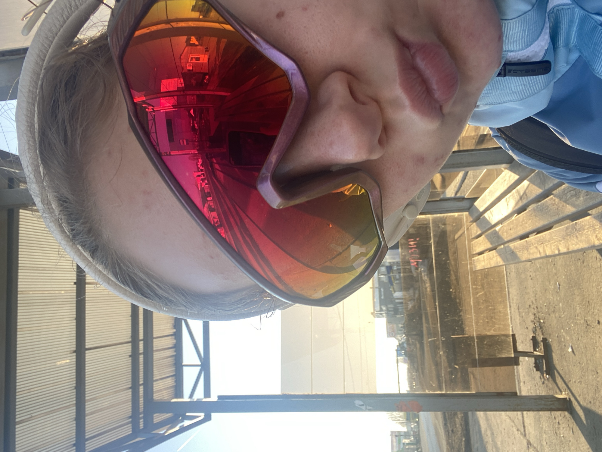 A person wearing sunglasses is positioned in front of a window, with an industrial or construction site visible outside. The reflection in the sunglasses shows a building with windows and structural elements. The person's face is partially visible, including the nose and lips. They are wearing a white cap or headband and a light blue jacket. The window has metal bars and a gray and white corrugated roof. The ground outside the window is a textured surface, possibly concrete or asphalt.