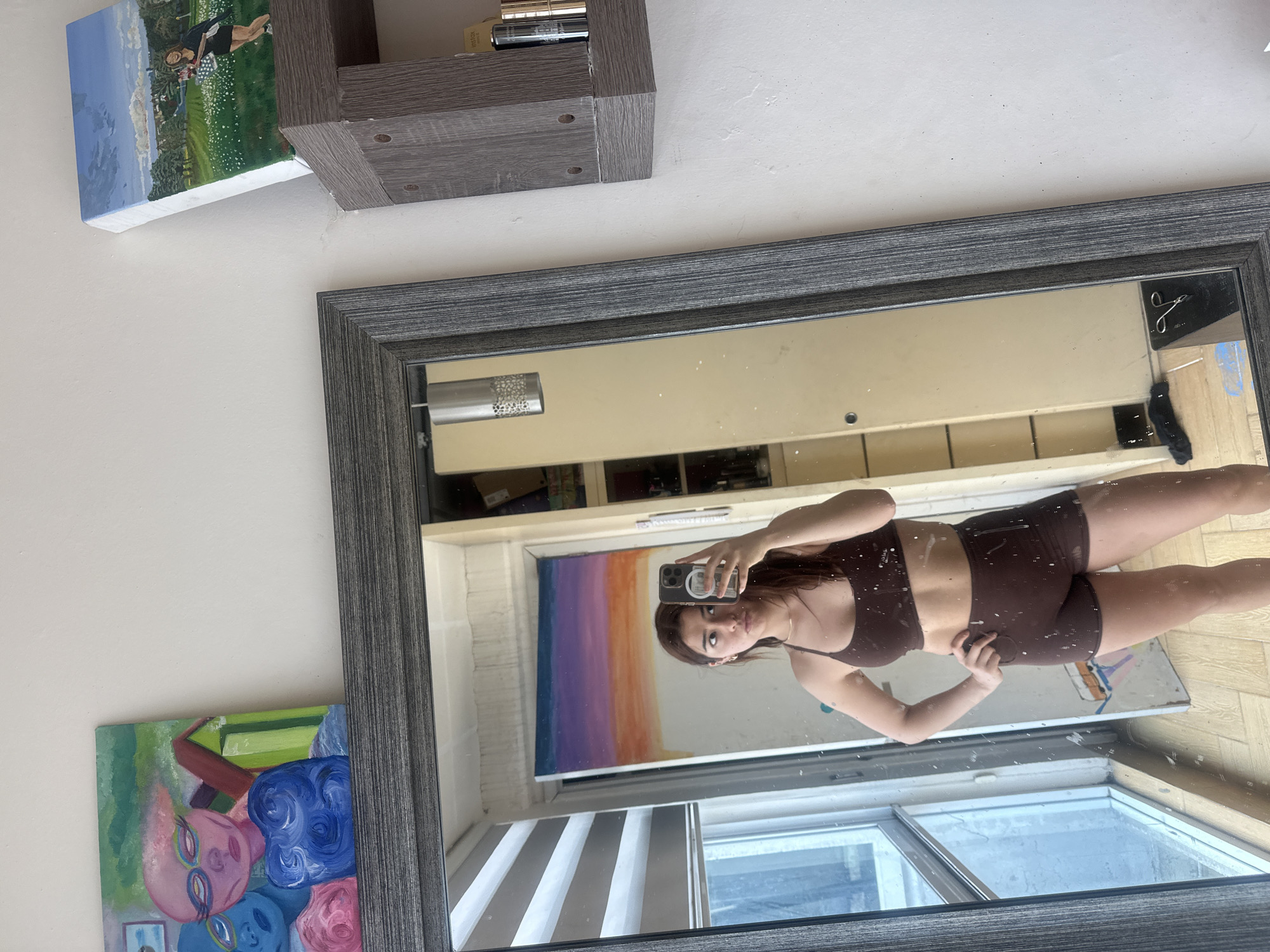A person is taking a selfie in a mirror. The person appears to be wearing a brown sports bra and shorts. Behind the person in the mirror is a room with an abstract painting on the wall. There is a window with blinds partially open. On the wall to the left of the mirror is a painting and a shelf. The location appears to be an indoor setting, but without additional context, it is not possible to definitively identify the city as Puebla, Mexico.