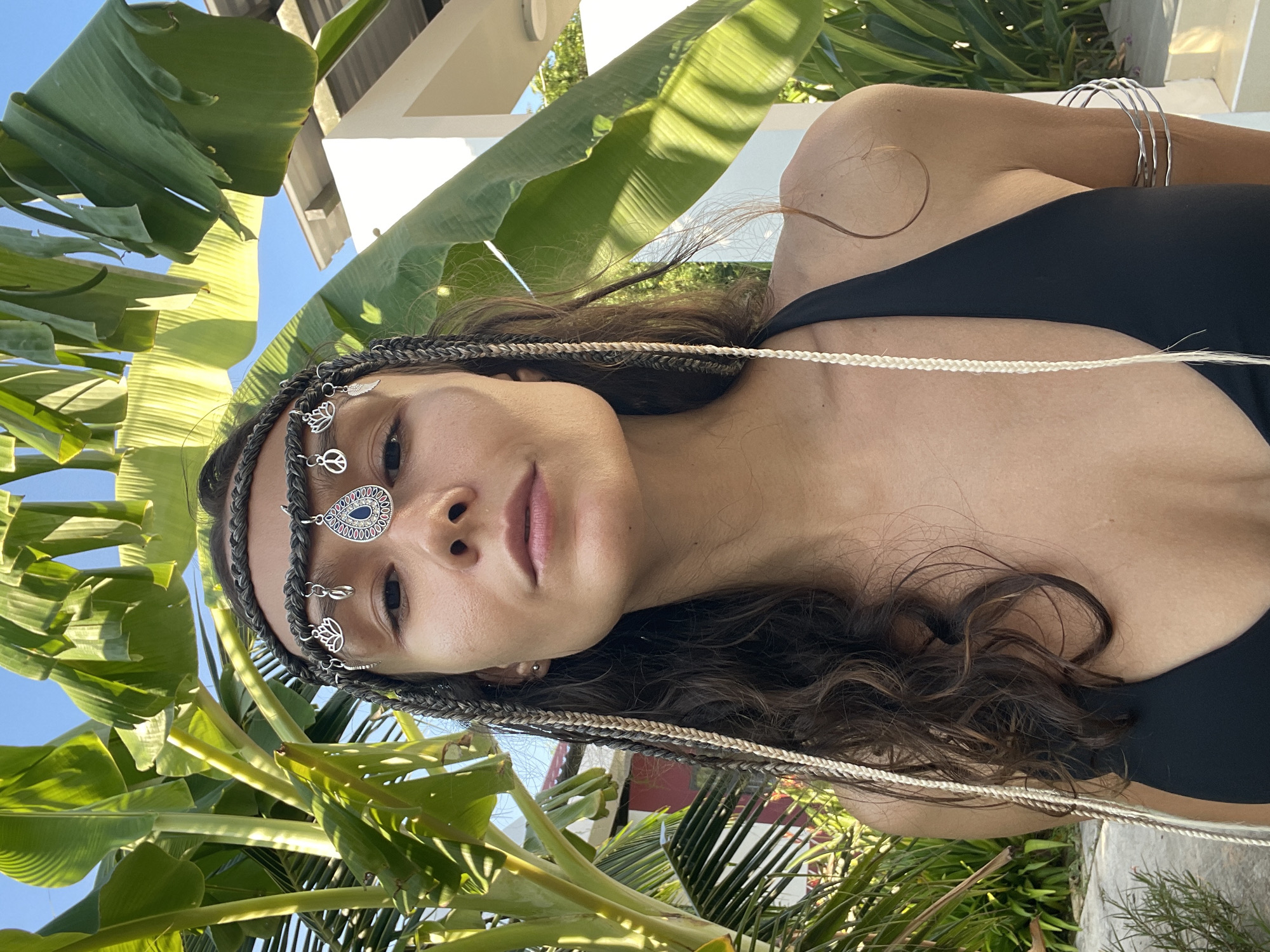 A medium shot captures a woman with braided hair, adorned with a decorative headband featuring charms like lotus flowers, peace signs, and a teardrop-shaped blue stone. She wears a black swimsuit and silver armbands. The background is filled with lush green banana leaves and a bright blue sky, with a glimpse of white modern architecture. The scene suggests an outdoor setting, possibly in a tropical or subtropical climate, such as Phu Thuy Ward, Vietnam, implied by the presence of banana trees and tropical foliage. The lighting indicates a sunny day, casting shadows and highlighting the subject.