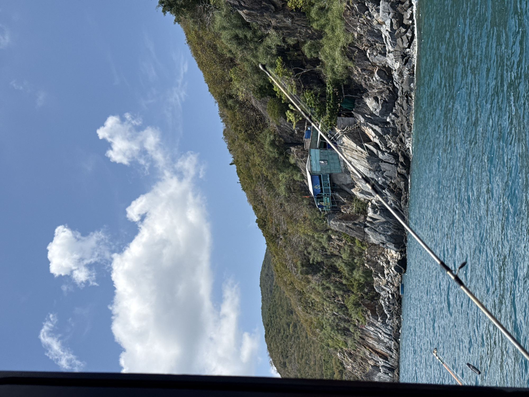 A slightly rotated view shows a rocky cliff face, likely in or near Bac Nha Trang Ward, Vietnam, as indicated. The cliff is covered in green vegetation and transitions into a body of water to the right. A small, turquoise-colored structure is perched on the cliff, accessible by a series of wooden steps or a walkway. Two fishing rods are visible in the foreground, partially obstructing the view. The sky above features a few scattered clouds. The bottom edge of the frame contains a dark, out-of-focus area, likely from the boat or structure the image was taken from. The focus is on the landscape and the small building on the cliff.