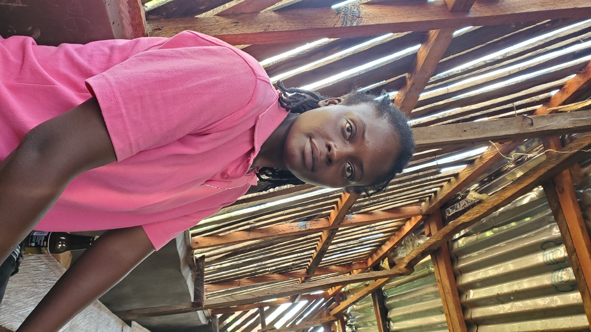 In the city of Jalingo, Nigeria, a medium shot captures a Black woman with dark skin and braided hair, wearing a pink polo shirt. She is seated, her arms resting on a table. The background consists of a rustic wooden structure with corrugated metal roofing. The woman's expression is neutral as she looks towards the camera. A dark-colored bottle is visible on the table to her right. The environment appears to be an outdoor setting, possibly a market stall or an open-air shelter, given the construction style and natural light filtering through the wooden slats.