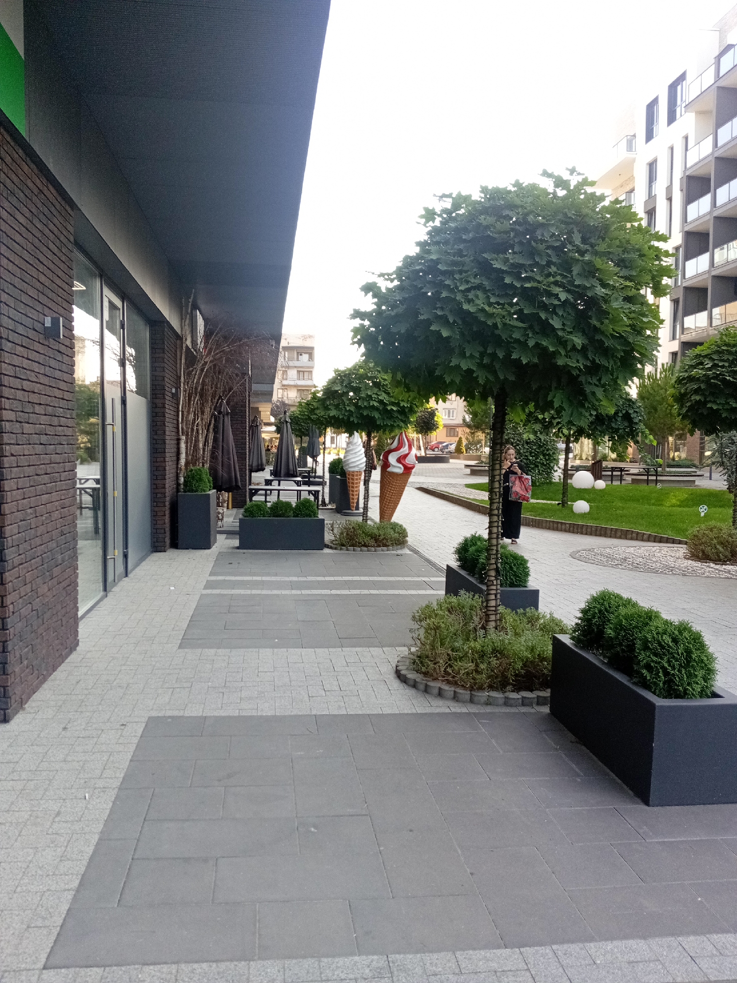 This is an outdoor scene in the city of Tiachiv, Ukraine, featuring a modern commercial building with a brick facade and large glass windows. Adjacent to the building is a paved walkway with a patterned design of gray and lighter gray stone tiles. Along the walkway are dark gray planters containing neatly trimmed green bushes. Two large decorative ice cream cone sculptures are visible, one with red and white swirls and another in white. Several trees with round canopies are planted in the vicinity, some with decorative trunk wrap. In the background, a contemporary apartment building with multiple balconies can be seen. A woman in a black dress and carrying a red bag is walking on the paved path, and several dark umbrellas are set up on tables in an outdoor seating area. The overall environment appears to be a well-maintained public space, possibly a plaza or courtyard outside a cafe or shop.