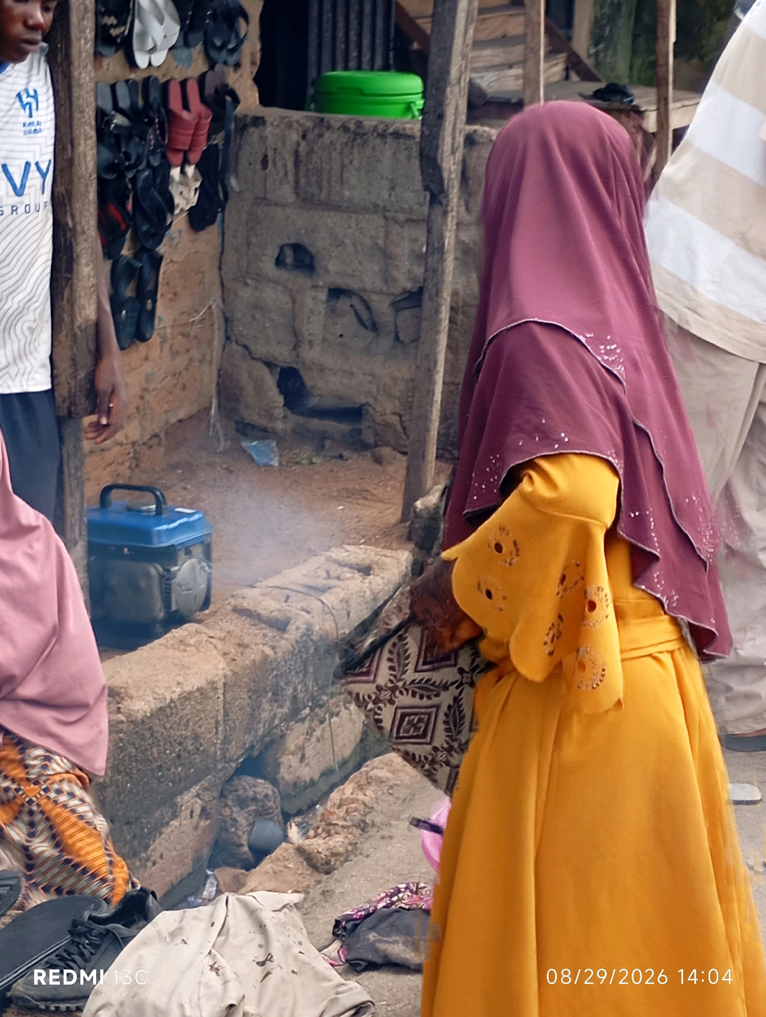 In Shendam, Nigeria, this image captures a street scene with a focus on a small generator emitting smoke. To the left, a person wearing a white t-shirt with blue accents and dark trousers stands next to a wooden post. A woman in the foreground is dressed in a vibrant yellow dress and a maroon hijab adorned with white embellishments. Her yellow sleeve features decorative cutouts. The background shows a wall constructed of rough stones and mud bricks, with various pairs of footwear hanging on the wall, suggesting a marketplace or informal shop. A bright green container is visible on top of the wall. The ground is covered in sand and debris. The generator, a blue portable unit, is positioned on a raised stone platform. In the lower right corner, a timestamp reads "08/29/2026 14:04."
