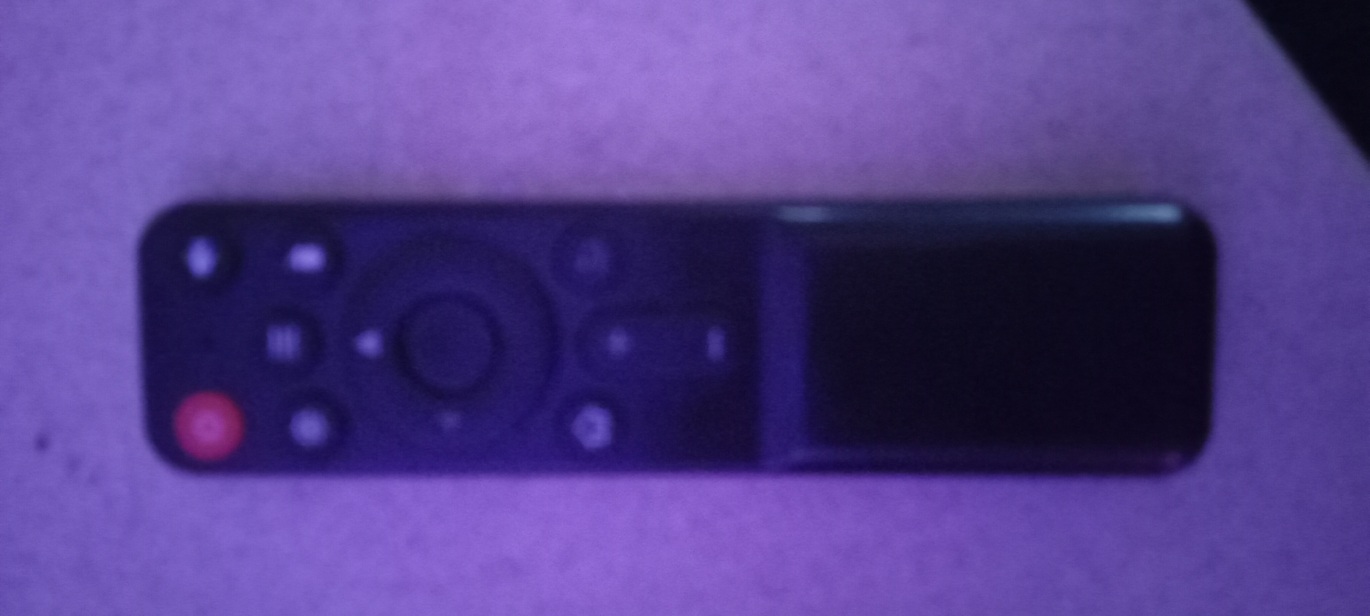 The image shows a black remote control with several buttons. The top button is red and appears to be a power button. Below that are several circular buttons, some with symbols. The bottom portion of the remote is a solid black rectangular area. The remote is placed on a textured surface that appears to be a light-colored fabric or wall, illuminated by purple light. The image quality is low, with significant blurriness. There is no information about the city of Perekip, Ukraine, in this image.