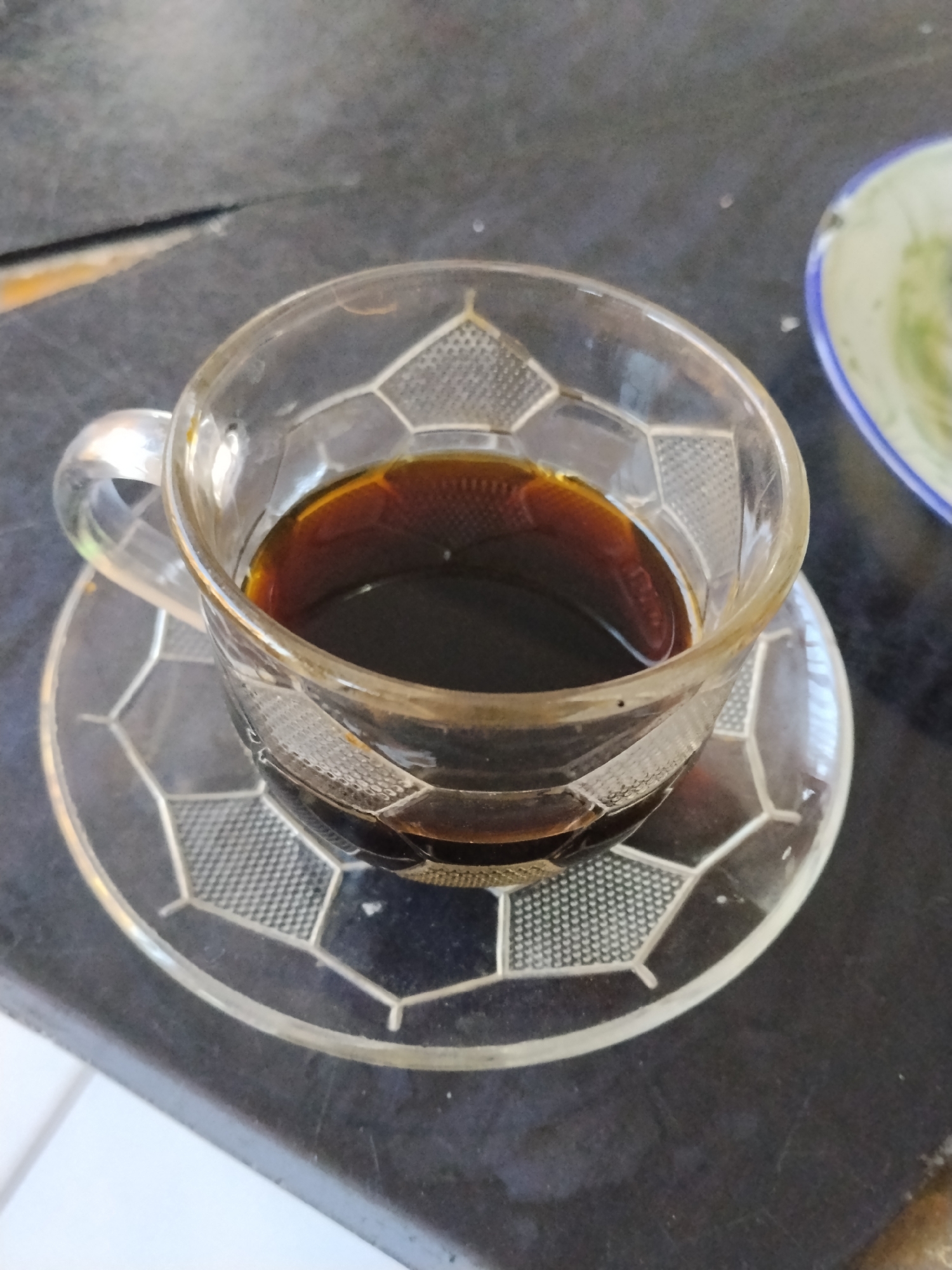 A close-up, overhead shot shows a clear glass coffee cup filled with dark liquid, sitting on a matching glass saucer. The cup and saucer have a raised, geometric pattern. The coffee cup is positioned on a dark, textured surface, possibly a table. To the right of the cup, a portion of a light-colored plate with a blue rim is visible. The background is out of focus, showing a dark, possibly wooden surface. The lighting is natural and casts subtle reflections on the glass. No people are present in the image. The setting appears to be an indoor environment.