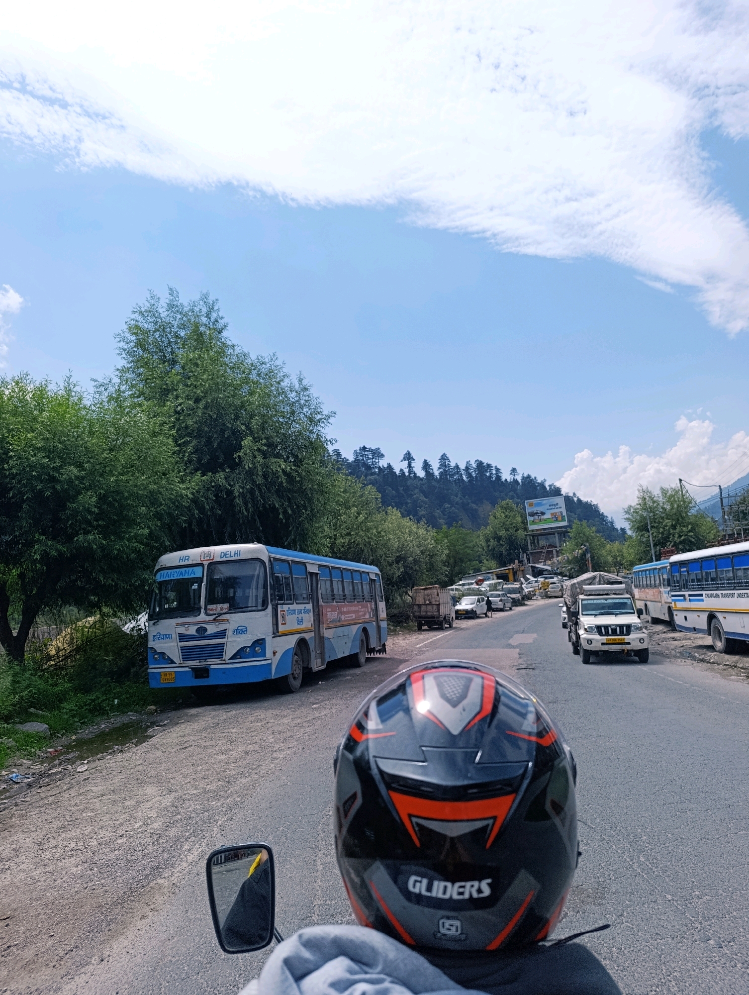 The image depicts a view from the perspective of someone riding a motorcycle in the city of Kalath, India. The road is paved and lined with trees and a blue and white bus on the left side. Further down the road, several other vehicles, including cars and a white SUV, are visible. In the background, a lush green mountainous landscape under a blue sky with white clouds completes the scene. The rider is wearing a black and orange helmet with the brand "GLIDERS" visible. The bus appears to be a public transport vehicle, indicated by the markings "HR 55 GY3005" and "HARYANA" and "DELHI" on its front. The overall atmosphere suggests a scenic drive through a mountainous region.