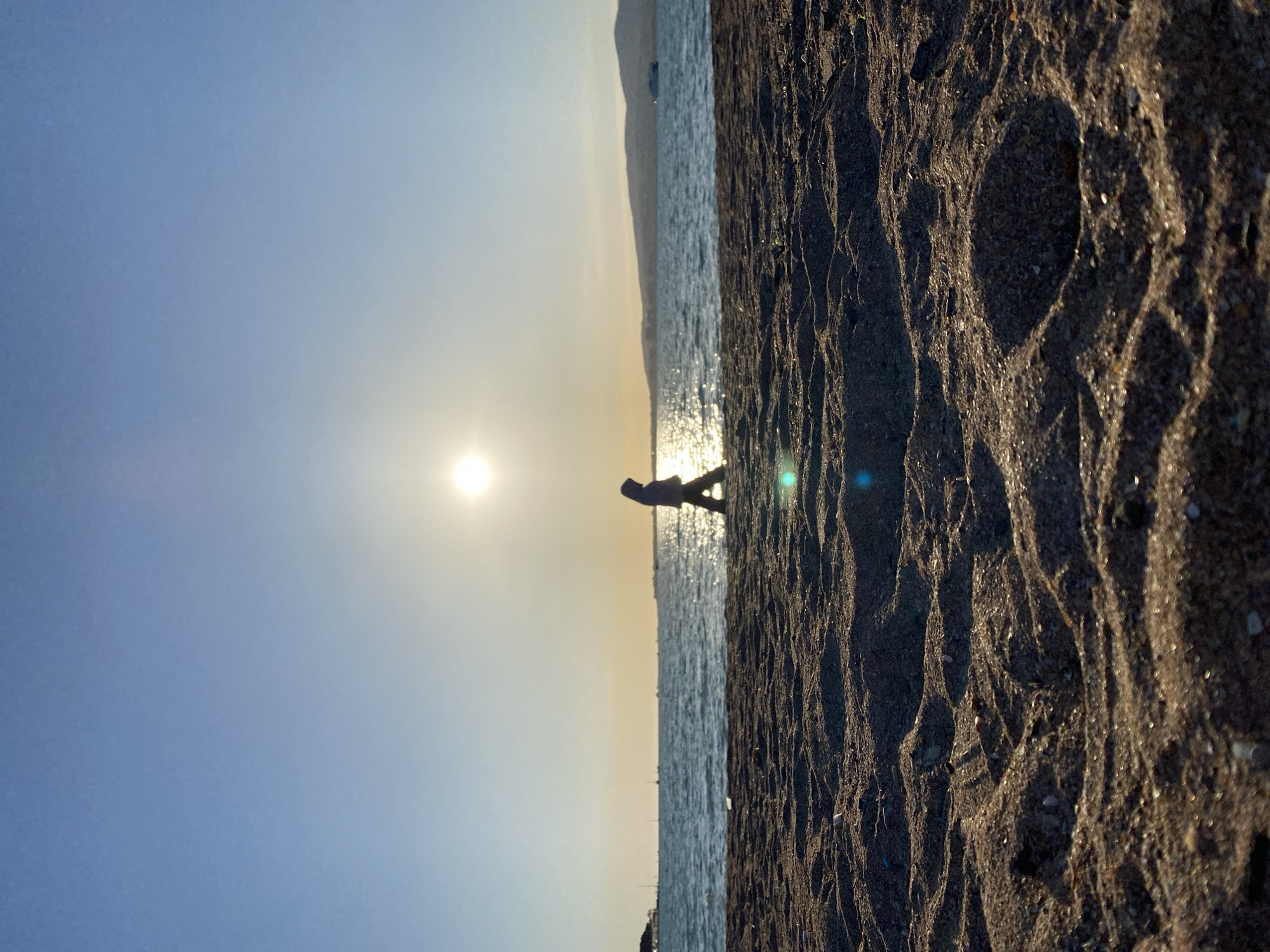 The image captures a serene beach scene at either sunrise or sunset in Gelendzhik, Russia. A solitary figure, rendered in silhouette, stands or walks along the water's edge, directly backlit by the bright, low sun. The sun's intense light creates a shimmering path across the calm sea, which stretches towards a distant, hazy mountain range visible as a dark silhouette on the horizon.
The foreground is dominated by dark, wet sand, heavily textured with ripples and small pebbles, suggesting the ebb and flow of the tide. The side lighting from the low sun accentuates these textures, creating defined shadows and highlights on the sand. The sky above is clear, displaying a gradient from a brilliant, warm glow near the sun to a softer blue further away. Several lens flares are visible in the bright areas of the image. The overall impression is one of peaceful solitude, set during the golden hour, with no visible text.