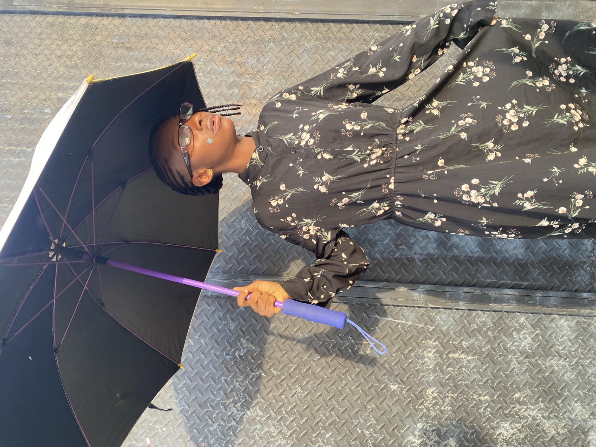 An individual is depicted holding a black umbrella with a purple shaft and ribs, featuring a blue foam grip and a light blue lanyard. The person, a young female with dark skin, wears a black, long-sleeved floral dress with light-colored blossoms and green leaves. She has braided hair, eyeglasses with reddish-brown frames, and a light blue star sticker on her left cheek. Her lips are slightly parted, exhibiting a pale, possibly glossy, coating. She is looking towards her right. The background consists of a dark grey metal wall with a diamond plate pattern, showing areas of wear and discoloration. Strong, direct lighting from the upper left creates distinct shadows. The scene is located in Iranla, Nigeria.