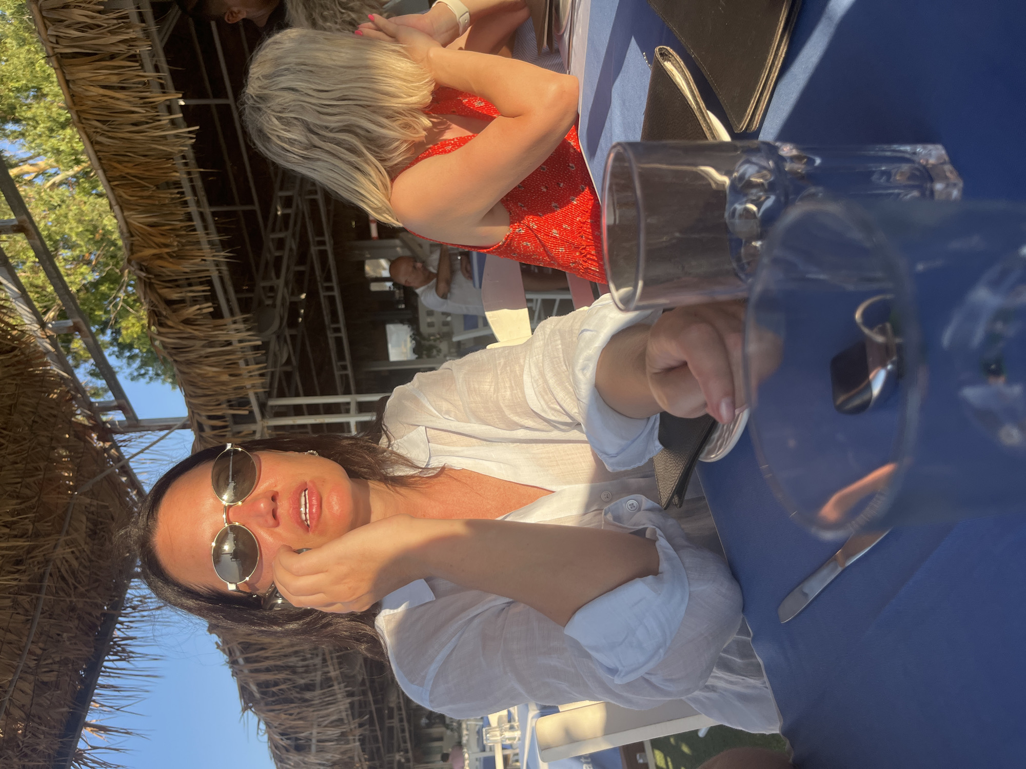 The image shows a woman sitting at a table outdoors, under a thatched roof structure. She is wearing sunglasses and a white shirt, and appears to be on the phone. In the background, another woman in a red dress is visible, and further in the background, a man is seated at a table. The setting appears to be a restaurant or cafe, likely near a beach, given the style of the overhead covering and the presence of outdoor seating. The lighting suggests it is either early morning or late afternoon. The location is specified as Polis Chrysochous, Cyprus.