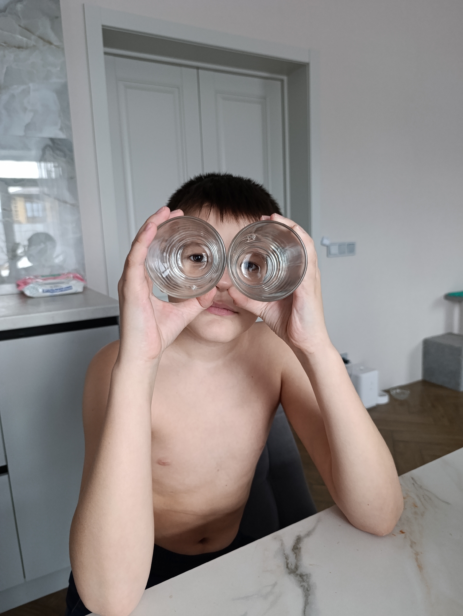 A shirtless, young boy is sitting at a table. He is holding two empty drinking glasses up to his eyes, as if they were binoculars. The background includes a kitchen counter with cabinets, a white door, and a glimpse of an outdoor scene. The table is white with a marble-like pattern. It is likely an interior shot taken in a residential setting, although a precise location in Chaikovsky, Russia is indeterminable without more specific details.