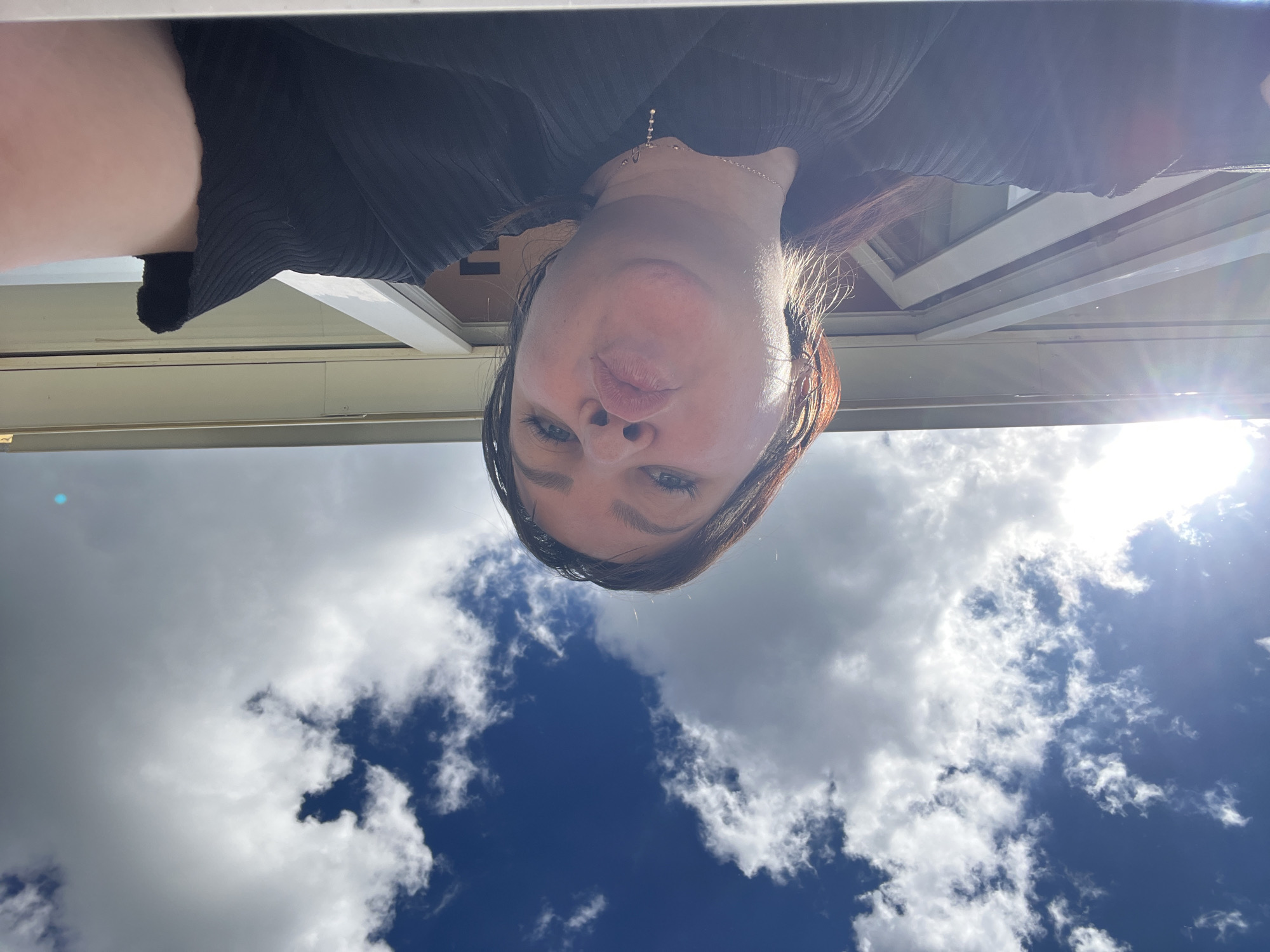 A selfie taken from a low angle. The subject is a woman with brown hair and blue eyes. She is wearing a black shirt and a necklace. The background is a blue sky with white clouds and a building. The location is Noviy, Russia.