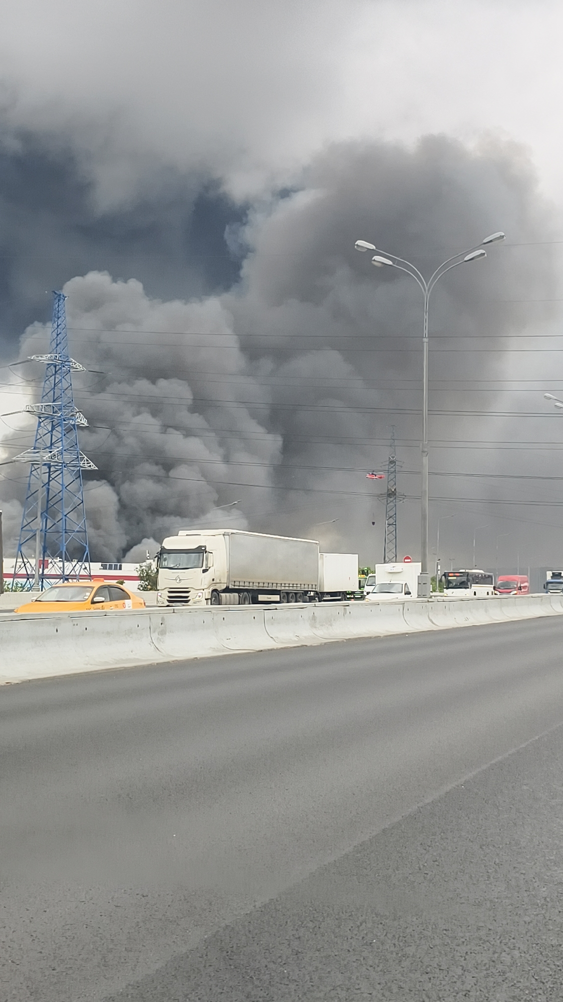 In the city of Unknown, Unknown, a plume of thick, dark smoke billows into the sky, obscuring the upper portions of industrial buildings and high-voltage power lines. A tall, blue lattice tower stands prominently on the left, with power lines stretching across the frame. In the foreground, a multi-lane highway is visible, separated by a concrete barrier. On the highway, a white semi-trailer truck with a full cargo trailer drives past, followed by other vehicles including a yellow taxi, a white van, and buses. The sky is overcast with a bright, hazy quality. The overall scene suggests a significant event, likely a fire, impacting the urban industrial landscape.