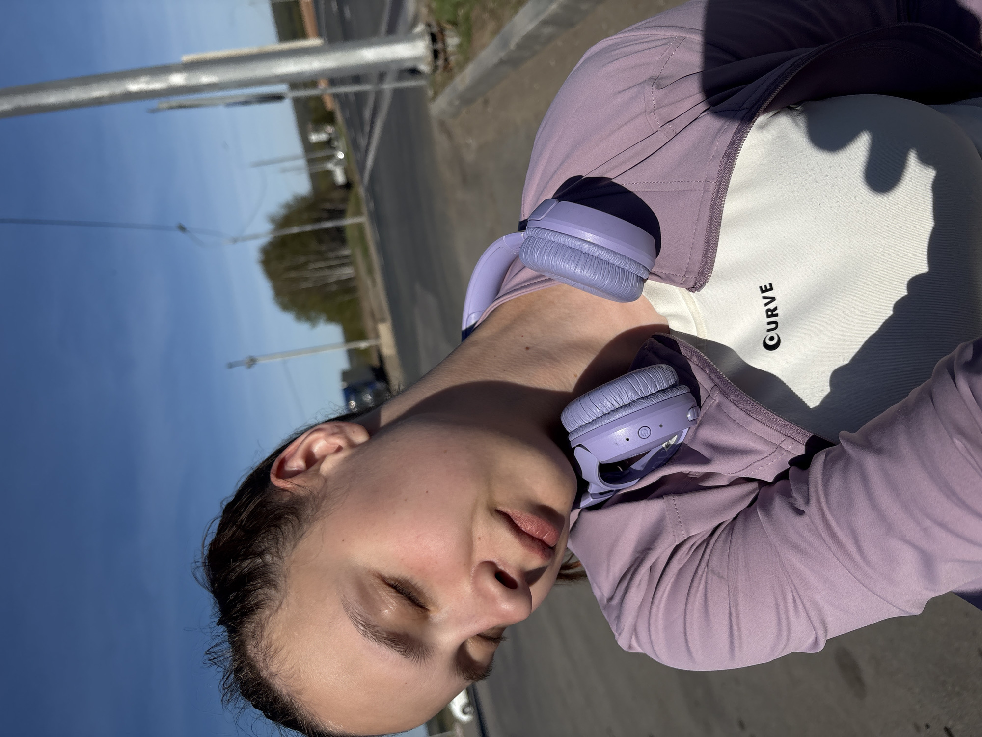 A woman stands outdoors in Verkh-Sosnova, Russia. She is wearing a light purple jacket over a white shirt with "CURVE" printed on it. Light purple headphones rest around her neck. The background features a road, trees, and a clear sky.