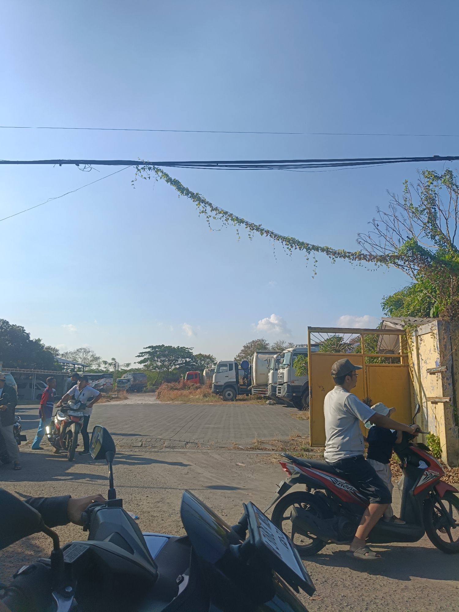 This is a medium shot, captured at eye level, of an outdoor scene on a bright, sunny day, likely in Kembangan, Indonesia. In the foreground, a portion of a black scooter is visible, with its handlebars and a hand gripping them. To the right of the foreground scooter, a red and black Honda scooter is prominently featured, ridden by a man wearing a grey t-shirt, black shorts, and a light-colored cap. He is facing forward, and a young child, wearing a dark shirt and a light-colored hat, is seated in front of him. The scooter is on a paved road.
In the mid-ground, several people are gathered. Two young boys, one in a red shirt and the other in a blue shirt, stand near a motorcycle. Further back, a group of men appear to be observing or interacting. A fleet of white trucks, some appearing to be concrete mixer trucks, are parked in the background. Beyond the trucks, trees and vegetation are visible, suggesting an area that might be industrial or on the outskirts of the city. A tall, weathered yellow gate stands on the right side of the mid-ground, adjacent to a building with a similar yellow hue. Overhead, power lines stretch across the frame, with a vine of green leaves trailing along one of them. The sky is clear and blue with a few scattered white clouds. The overall impression is a snapshot of everyday life in an Indonesian town.