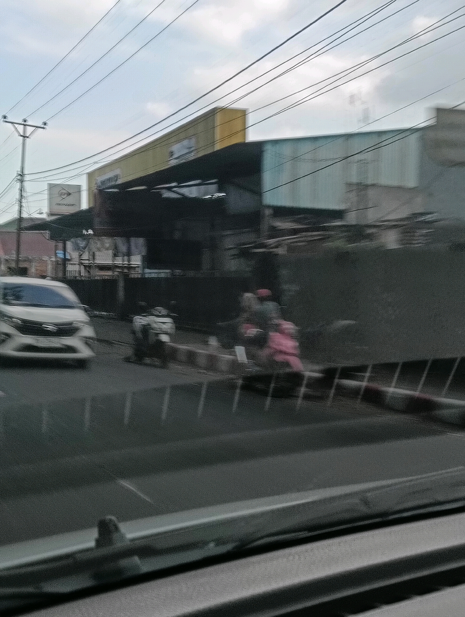 This is a motion-blurred, medium-angle, exterior shot of a street scene in Tomohon, Indonesia, likely taken from a vehicle. The image captures a cloudy sky overhead with multiple power lines crisscrossing the frame. In the background, a yellow building with a dark awning is visible, along with a light blue-gray building. A white car is in the left foreground, driving on a road. To the right of the car, a scooter is parked or moving slowly on the side of the road. Further to the right, two figures on a pink scooter are visible, also in motion. The foreground shows the interior of a vehicle, including a dashboard and windshield wiper.