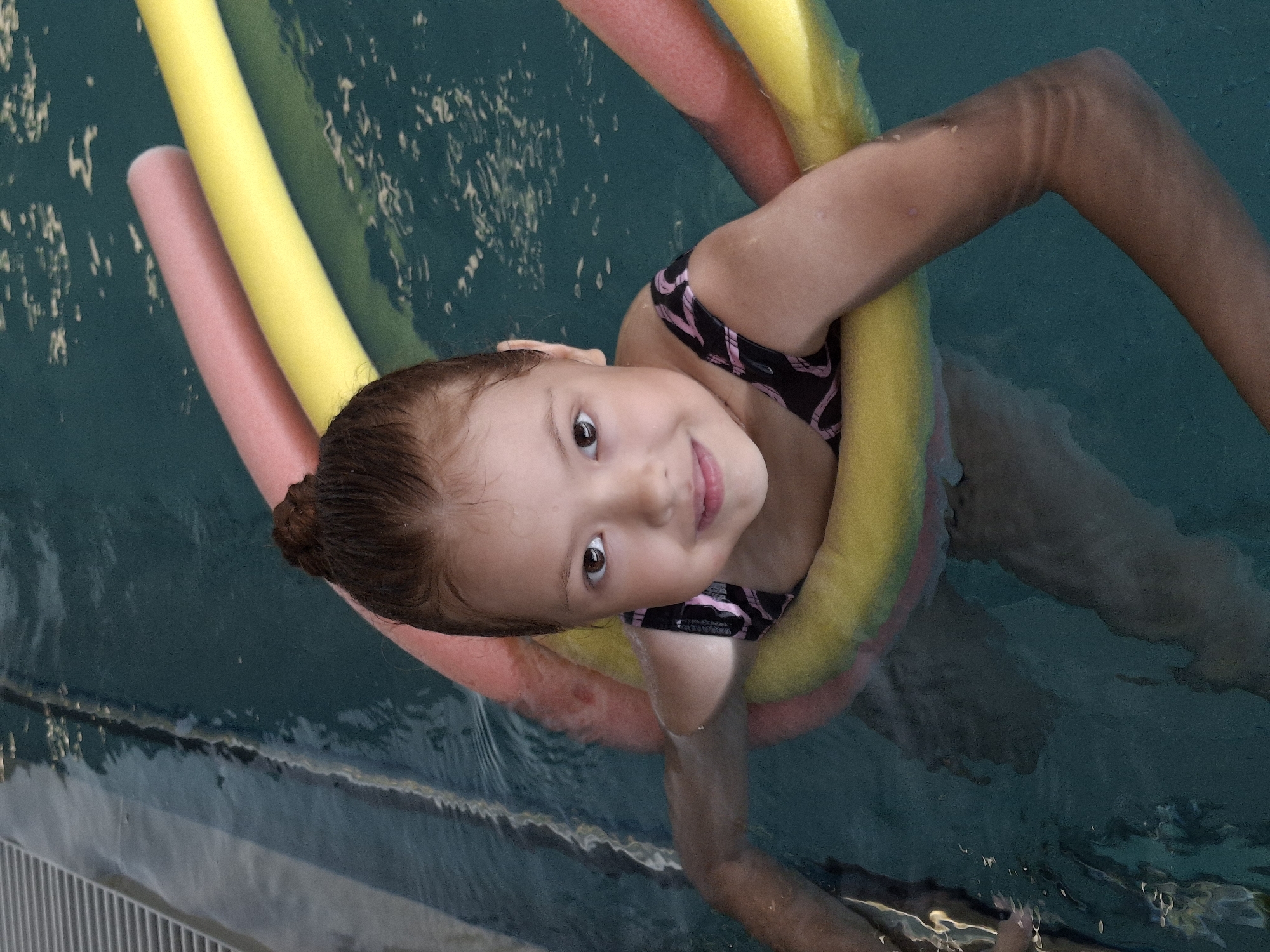 A young girl is swimming in a pool, supported by pool noodles. The noodles are yellow and pink, encircling her torso. She is wearing a black swimsuit with a pink pattern. The girl is looking up and smiling at the camera, with a happy expression. Her head is tilted to the side, and her hair is dark brown and pulled back. The water in the pool is a deep teal color, with reflections of light on the surface. The edge of the pool is visible at the bottom of the image, with a ribbed texture. The lighting suggests it is daytime, likely indoors or on an overcast day, as there are no strong shadows. The overall scene conveys a sense of fun and childhood enjoyment. There is no visible text in the image.