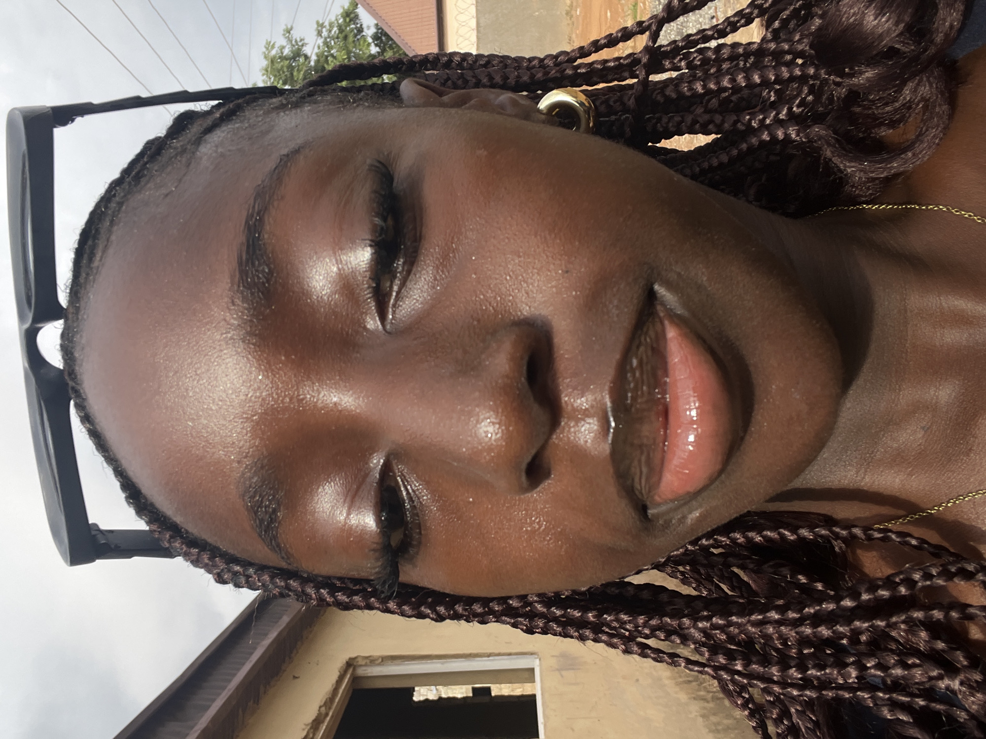 A close-up, tilted shot of a young, dark-skinned woman with brown braided hair. She is wearing a gold hoop earring in her left ear and a gold necklace. A pair of black sunglasses is resting on her head. Her eyes are closed, and her lips are slightly parted. The background includes a building with an open window and a roof. The location is possibly in Abuja, Nigeria, based on the image's origin.
