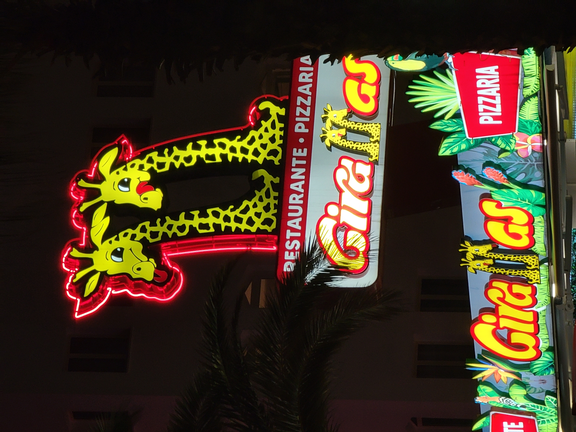 A neon sign for "Girafas" restaurant and pizzeria is displayed at night in Quarteira, Portugal. The sign features two cartoon giraffes made of red and yellow neon, with red outlining. Below the giraffes, the words "RESTAURANTE • PIZZARIA" are written in red lettering on a white background. Further down, the restaurant's name "Girafas" is prominently displayed in large, yellow, cursive neon letters, with the giraffe figures incorporated into the lettering. To the right, a red sign reads "PIZZARIA" in white lettering. The sign is set against a backdrop of palm trees and buildings, with the overall lighting from the neon signs illuminating the scene.