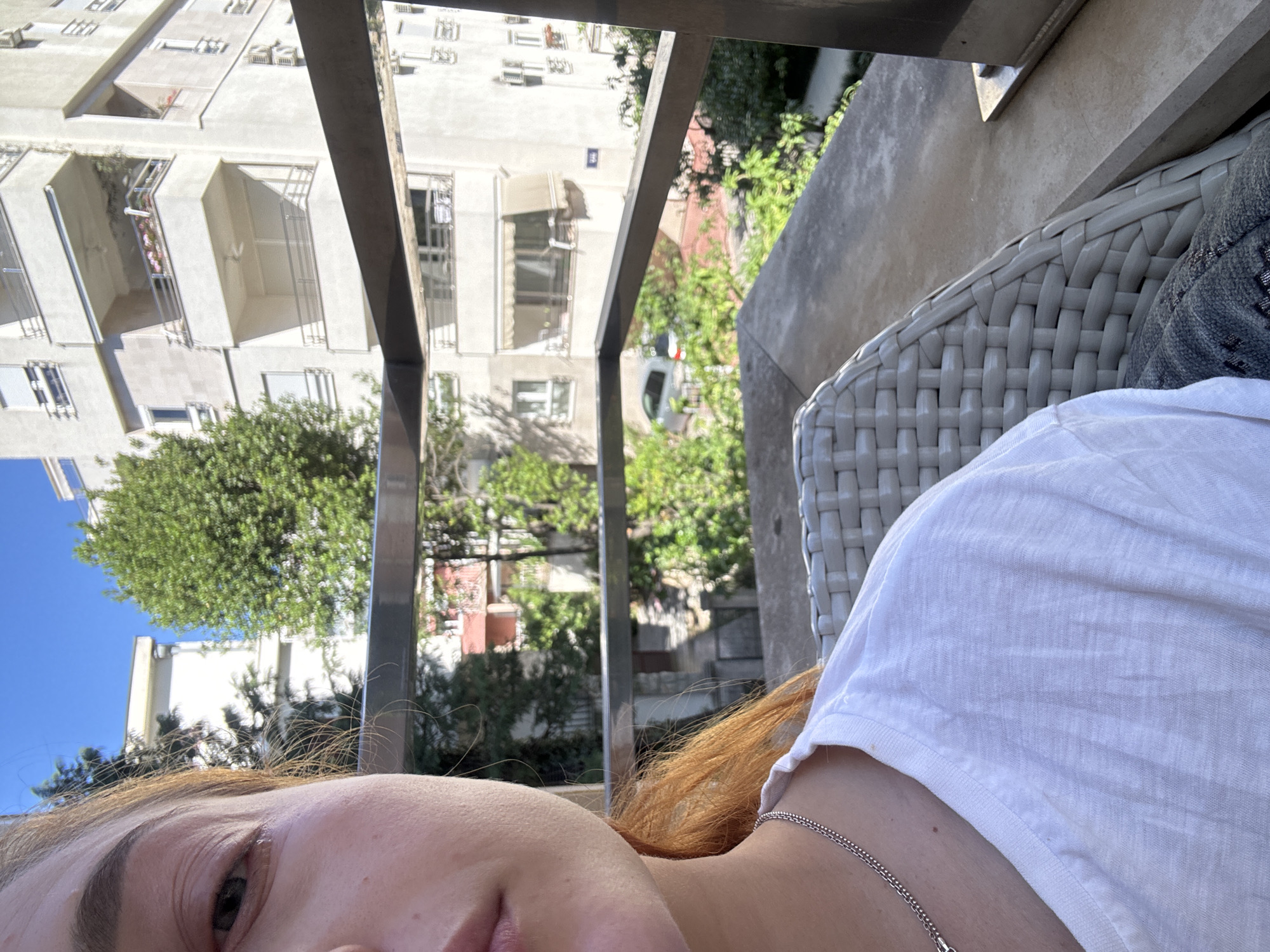 A close-up, tilted selfie shot shows the face and upper torso of a fair-skinned person with reddish-blonde hair, wearing a white t-shirt and a silver chain necklace. The person is partially visible from the left side of the image, with the left eye and part of the nose and mouth in focus. The background is visible through a metal balcony railing, presenting a daytime urban scene with a clear blue sky. A multi-story building with white facades and rectangular balconies is seen in the distance, partially obscured by green trees. The perspective is from an elevated position, suggesting the person is on a higher floor. To the right of the person, a white wicker chair with a grey cushion is partially visible. Based on the architecture, vegetation, and urban layout, this image may be taken in Budva, Montenegro, characterized by similar features.
