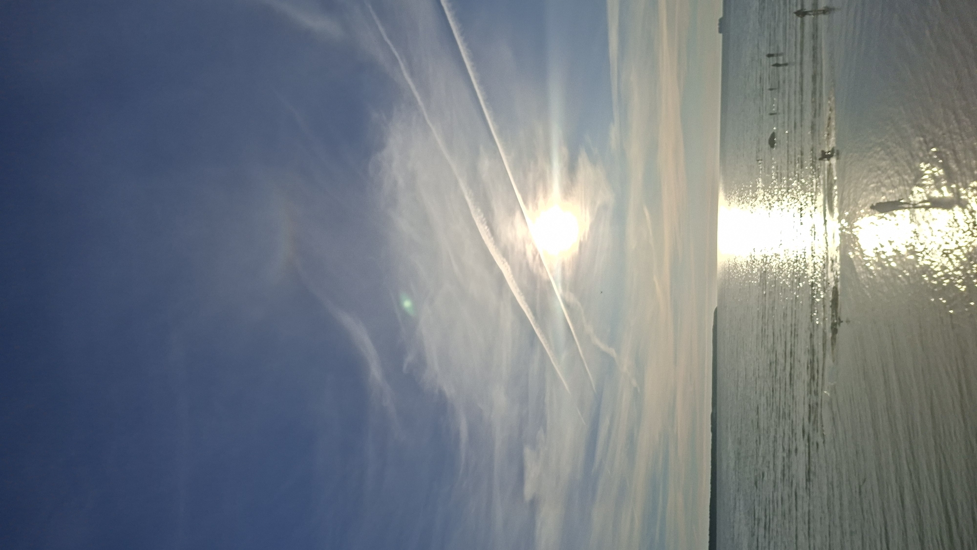 The image depicts a serene coastal scene with a vast expanse of calm, rippling water under a wide blue sky dotted with wispy white clouds and contrails from airplanes. The sun, a brilliant orb, is low in the sky, casting a shimmering golden reflection on the water's surface. Several small figures are visible in the shallow water near the shore, some appearing to walk or stand, and others possibly engaged in activities. A long, dark, tree-lined strip of land forms the horizon in the distance. In the far right of the horizon, a distant ship can be discerned. The scene suggests a peaceful late afternoon or early evening at a beach.
The location appears to be a coastal area in Tallinn, Estonia, possibly Pärnu Beach, known for its shallow waters and long sandy stretches, or perhaps a similar coastline along the Baltic Sea in or near the city.