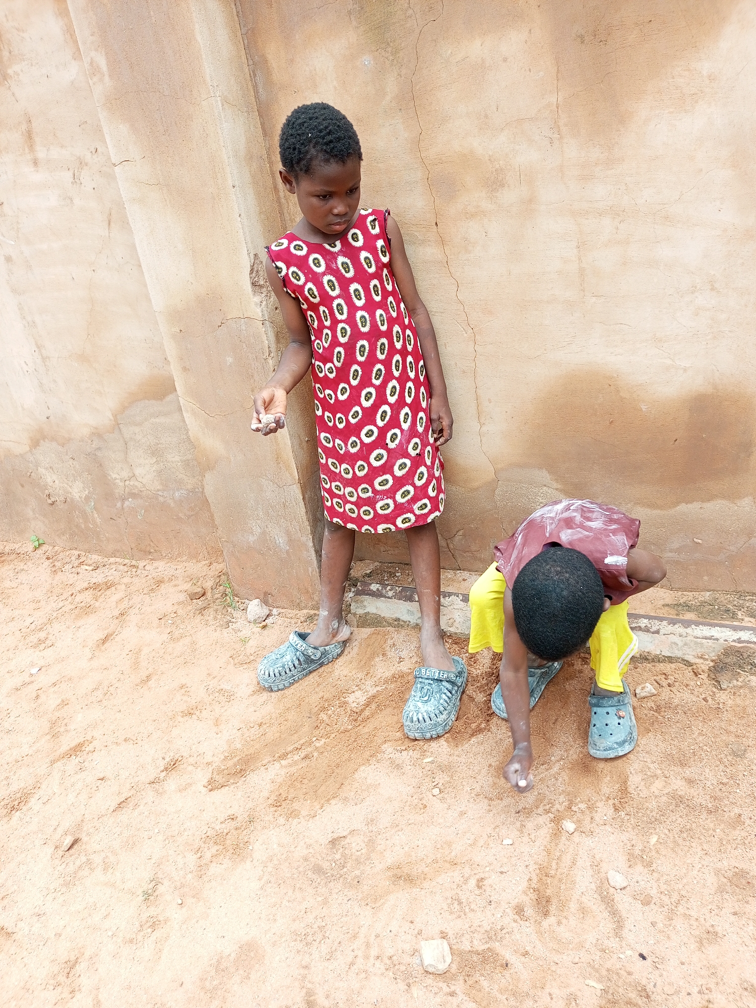 Two young children, one standing and one crouching, are in an outdoor environment with a sandy ground and a textured, light brown wall behind them. The standing child, a girl, wears a red dress with a white and black circular pattern and large, light blue clogs. She is holding small objects in her right hand. The crouching child, facing away from the camera, wears a maroon shirt and yellow shorts, also in large, light blue clogs. The child's left hand is reaching towards the sandy ground. The scene appears to be a simple, everyday setting, possibly in Tempure, Nigeria, given the architectural style of the wall and the sandy terrain, which are characteristic of many regions in Nigeria.
