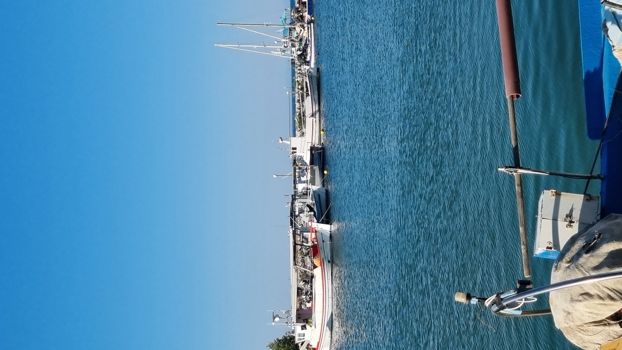 This is a vertical, eye-level, outdoor, daytime, medium shot photograph of a harbor in Limani Litochorou, Greece. The foreground shows the railing of a boat with a beige tarp and a white metal box with a blue lid. The middle ground is a body of blue water with several boats docked along a pier in the background. The boats are a mix of fishing vessels and sailboats, with white hulls and various colored accents. They are moored side-by-side. The background is a clear, bright blue sky. There are no people visible in the image. The overall impression is of a calm, sunny day at a Greek harbor.