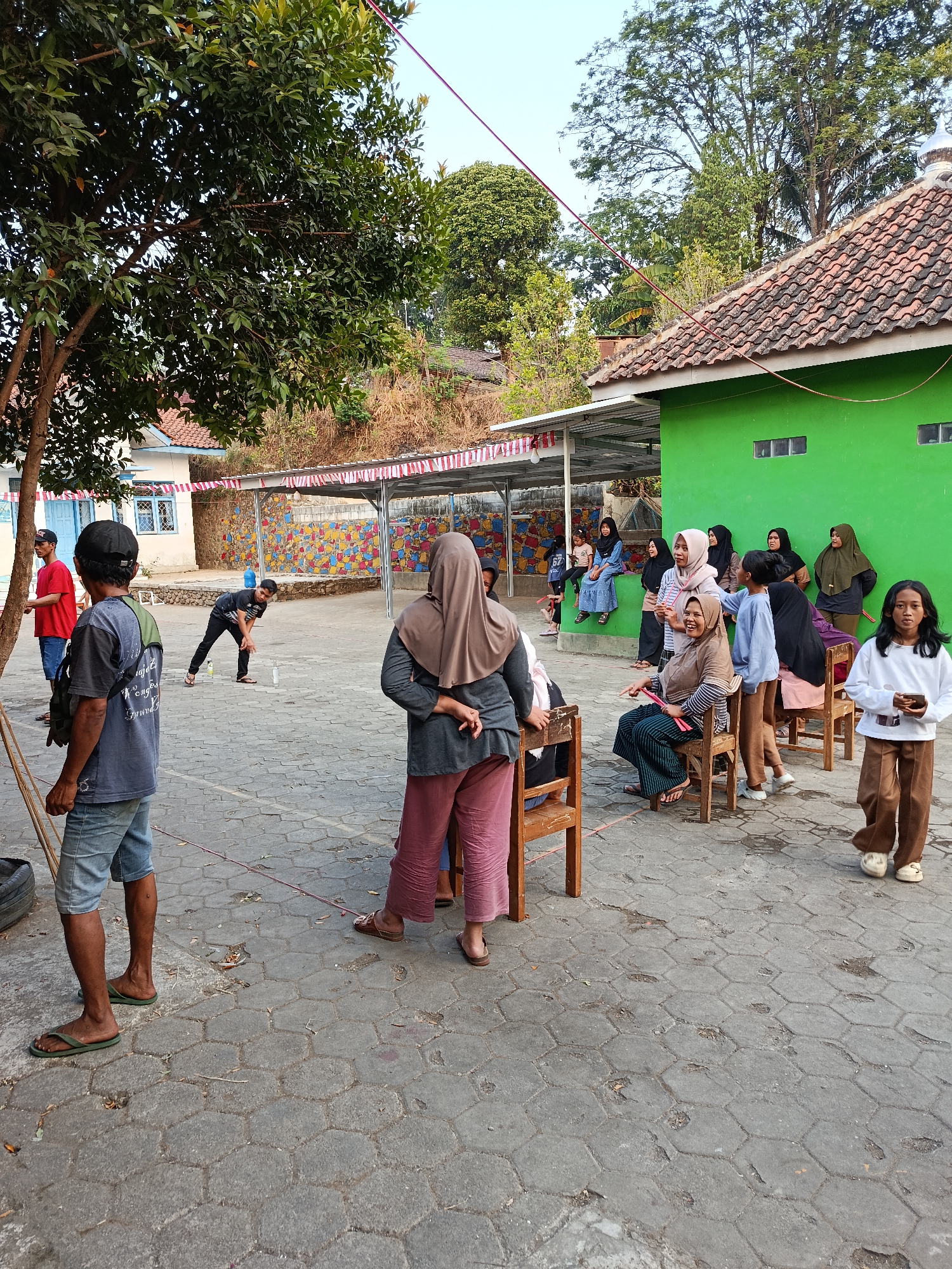 In Sidomukti, Indonesia, a group of people are gathered outdoors in a paved courtyard. A tree with green foliage stands to the left, casting shadows. In the background, a vibrant mural adorns a wall, and a bright green building with a tiled roof is visible. Several individuals, including women wearing hijabs and children, are seated on wooden chairs or standing. Some people appear to be engaged in a game or activity involving a bottle on the ground. The ground is paved with hexagonal stones. The overall atmosphere suggests a communal gathering or event.